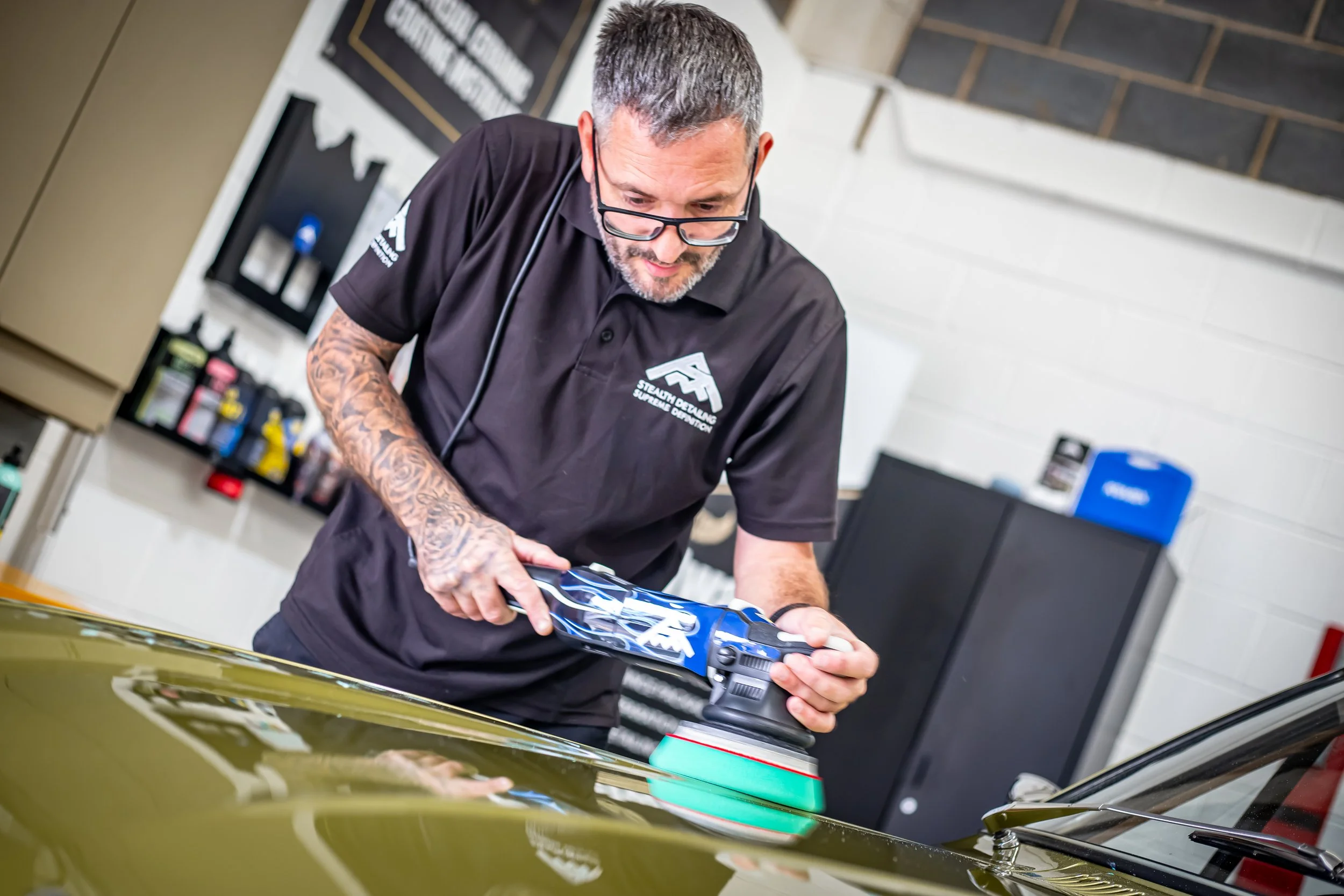 A man with glasses and tattoos on his arms applying a decal to a green car's hood at a car detailing shop.