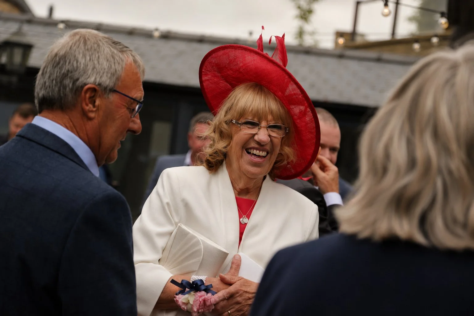 A woman wearing a large red hat and glasses is smiling and shaking hands with a man in a suit at an outdoor event, with others in the background.