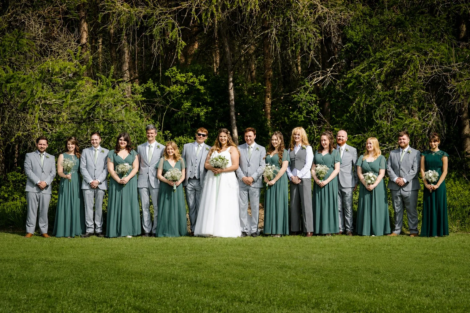 A wedding party standing outdoors on grass with trees in the background. The group includes bridesmaids in green dresses and groomsmen in gray suits, with the bride in a white wedding gown and the groom in a gray suit, all smiling.