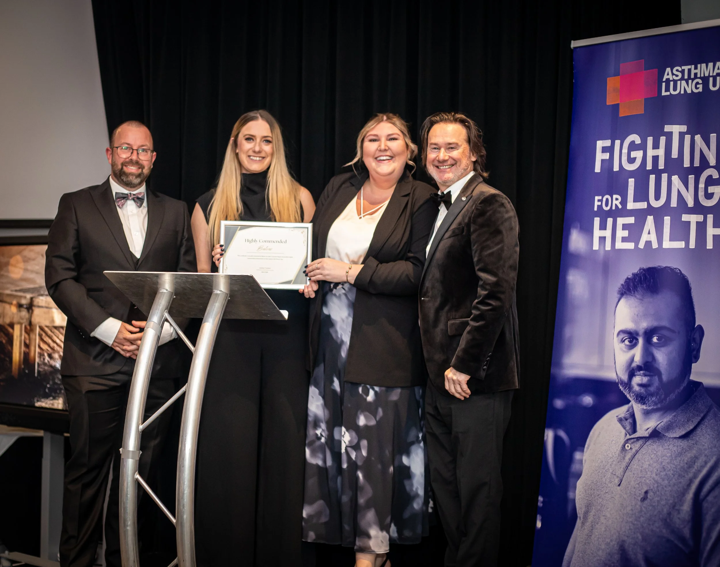 Group of four people on stage at an award ceremony, with a woman holding a framed certificate, standing next to a man in a tuxedo, and a woman in a blazer with a patterned skirt, and another man in a tuxedo with a bow tie. A banner to the right reads