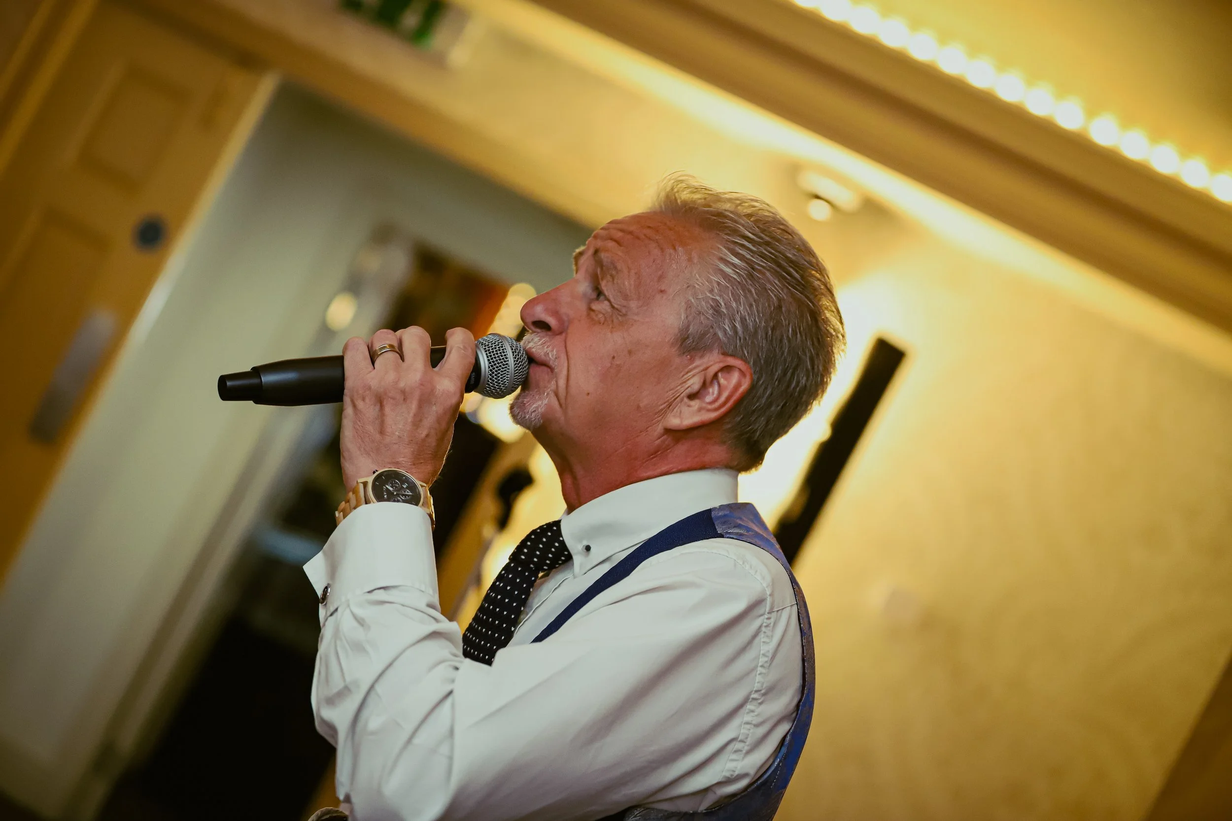 An older man singing into a microphone at an indoor event, dressed in a white shirt, black polka dot tie, and suspenders.