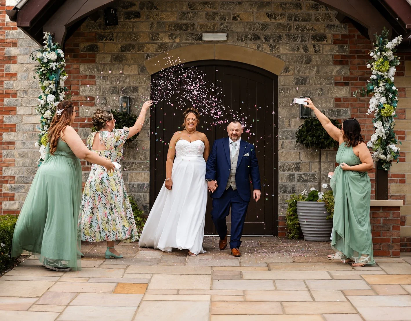 A newlywed couple happily walking out of a building as confetti falls around them, with two women on their sides taking photos and throwing confetti, and a floral arch framing the doorway.