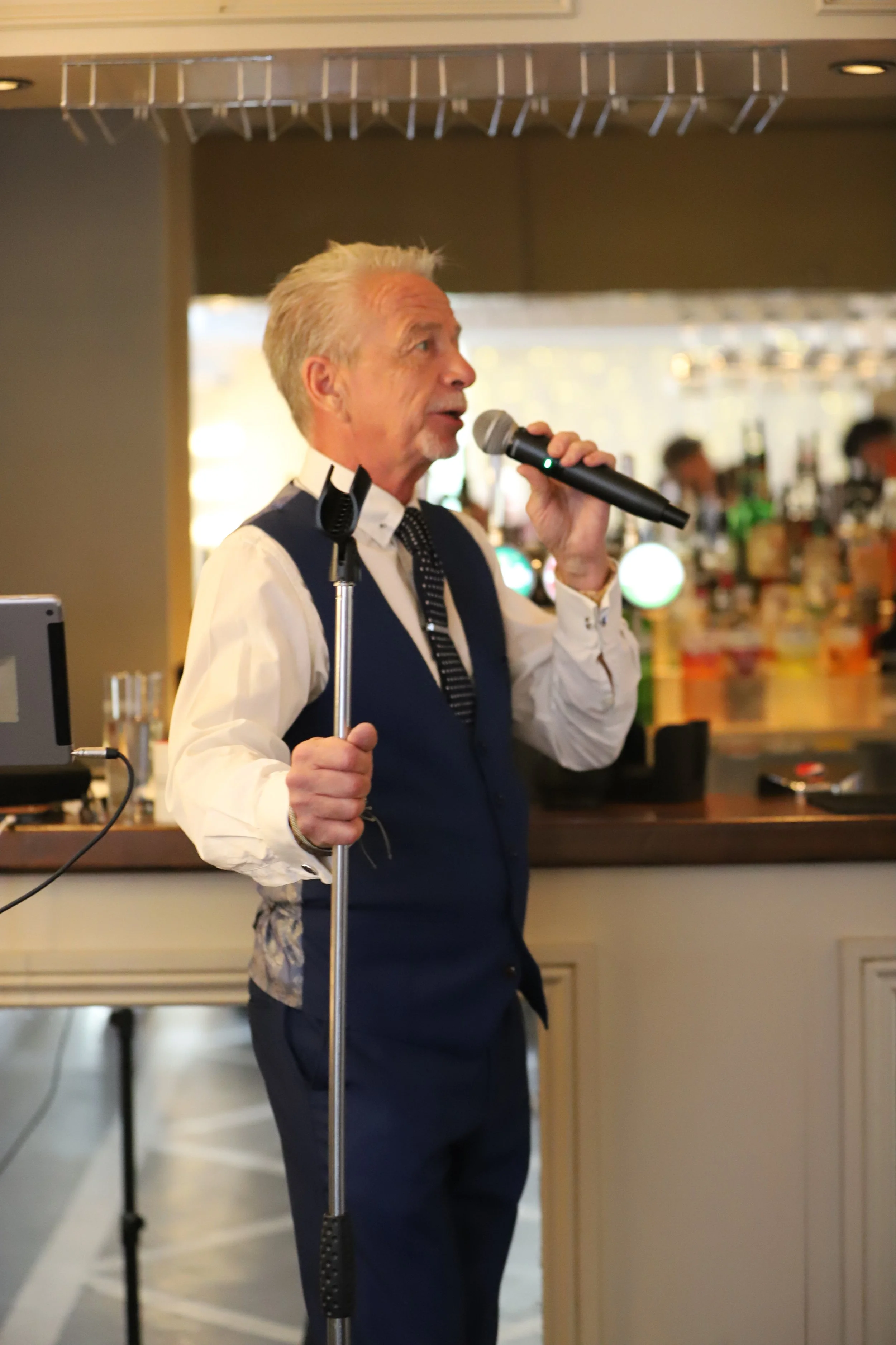 An elderly man with gray hair and a goatee, wearing a white shirt, navy vest, and polka-dot tie, is speaking into a microphone at a bar or restaurant with a bar counter and bottles in the background.