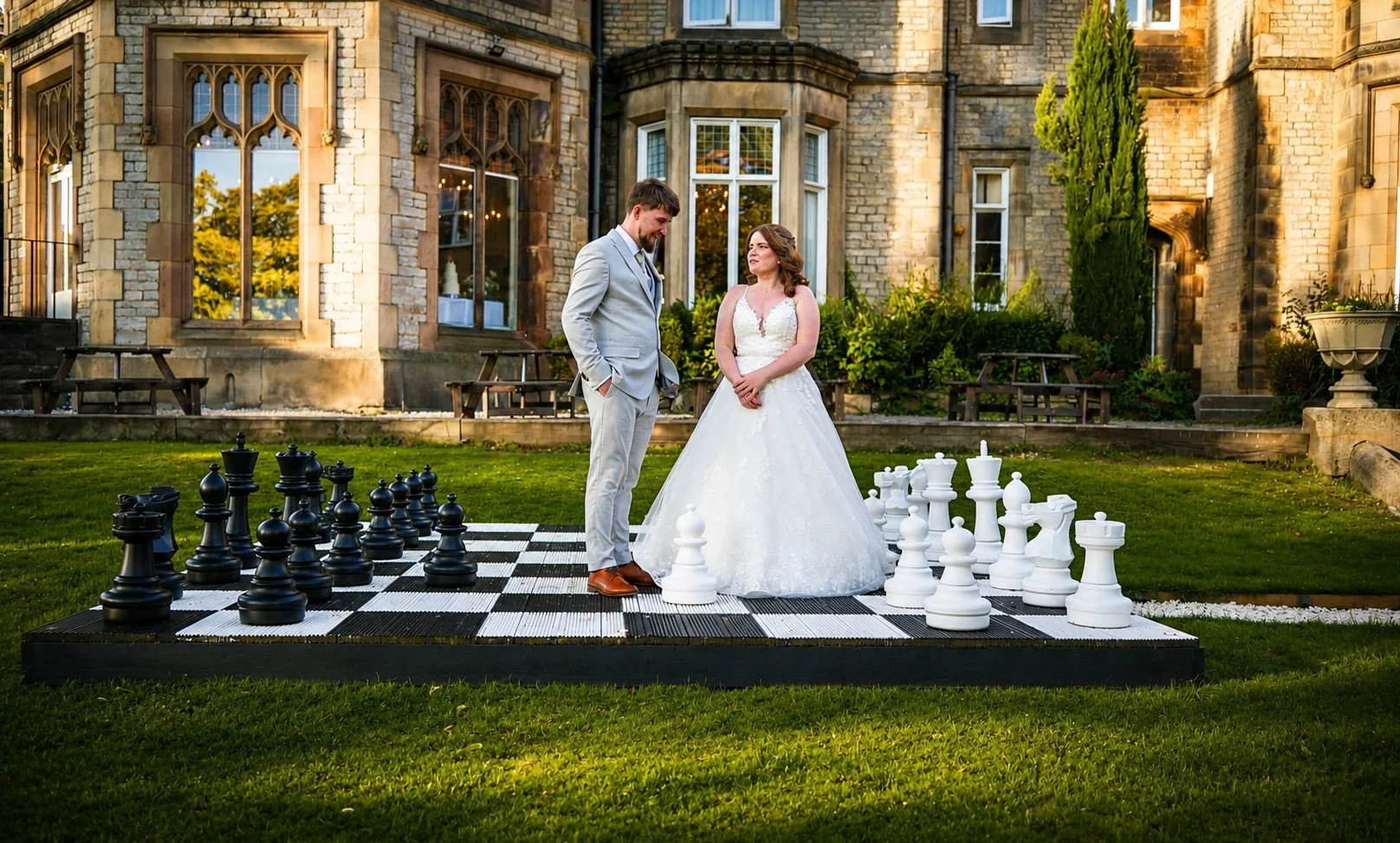 A bride and groom standing on a large outdoor chessboard with oversized chess pieces in front of a historic-looking stone building, during sunset.