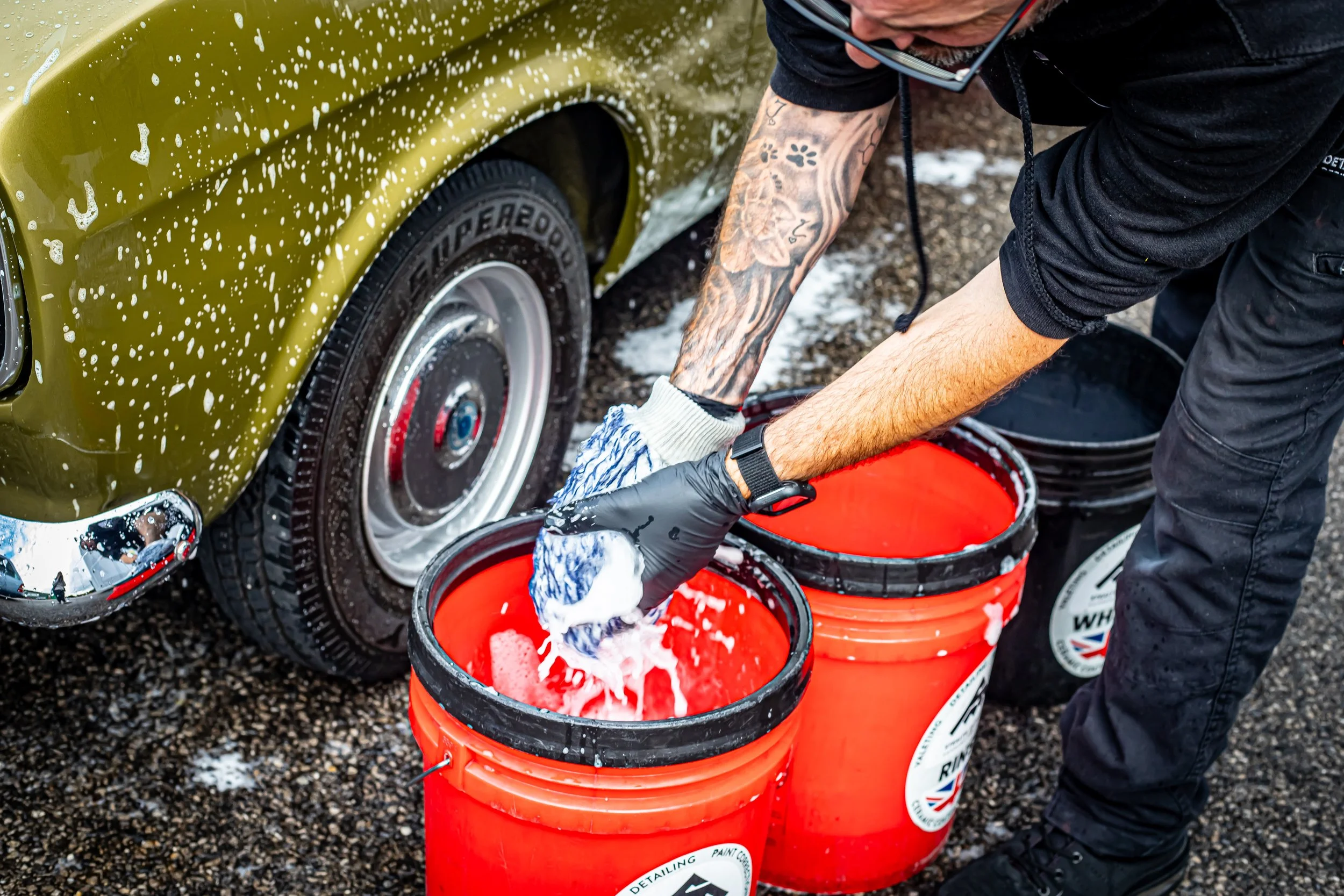 A person washing a green car with bubbles using their hands. They are wearing black gloves and have tattoos on their arms. There are multiple orange buckets filled with soapy water, and the person is leaning over the buckets while cleaning the car.