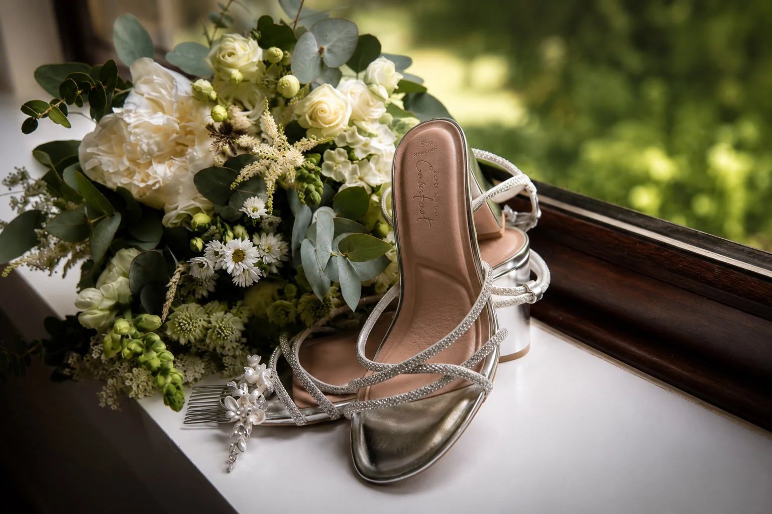 A pair of silver high-heeled shoes with rhinestone straps, a bridal hair comb with pearls, a white and green bridal bouquet, and a wooden window ledge with greenery outside.