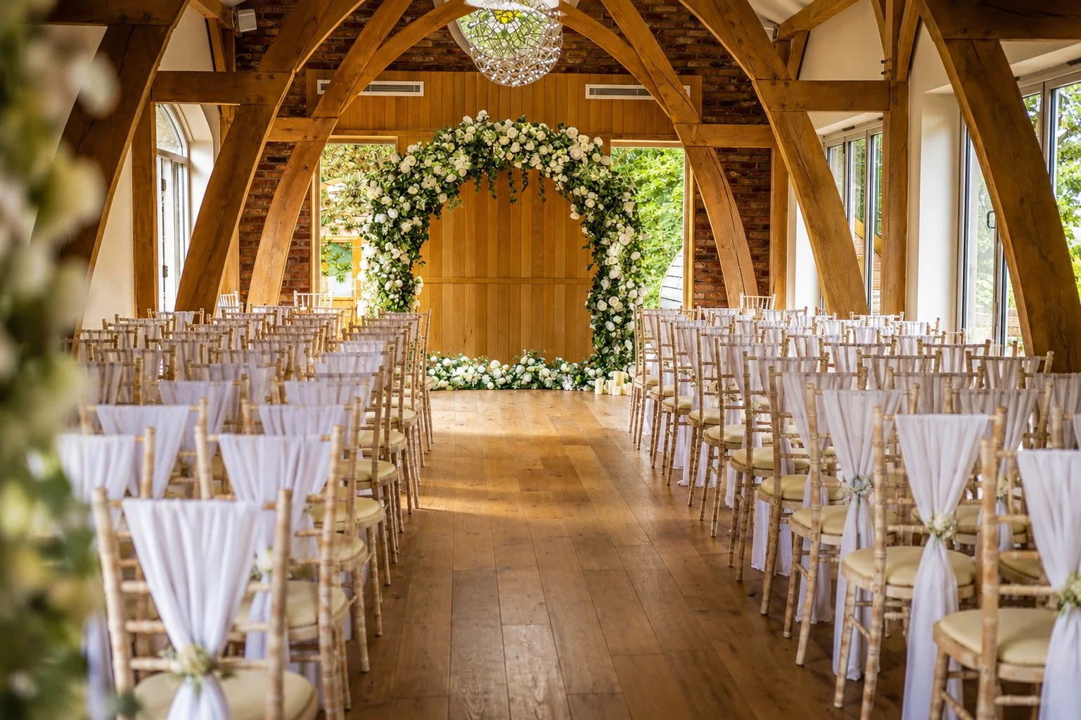 An indoor wedding ceremony setup with rows of wooden chairs decorated with white fabric and small flowers, facing a floral arch with white roses and greenery, inside a rustic wooden structure with large windows and an arched ceiling.
