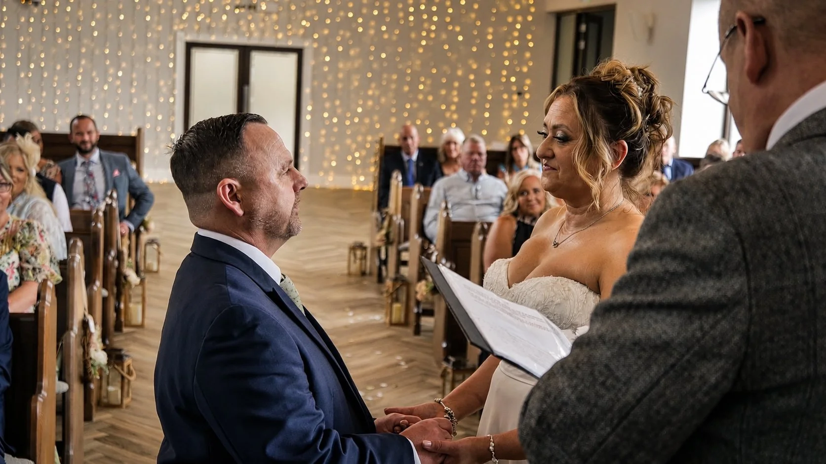 A wedding ceremony with a bride and groom holding hands and gazing into each other's eyes, standing before an officiant, in a decorated venue with guests seated in the background.