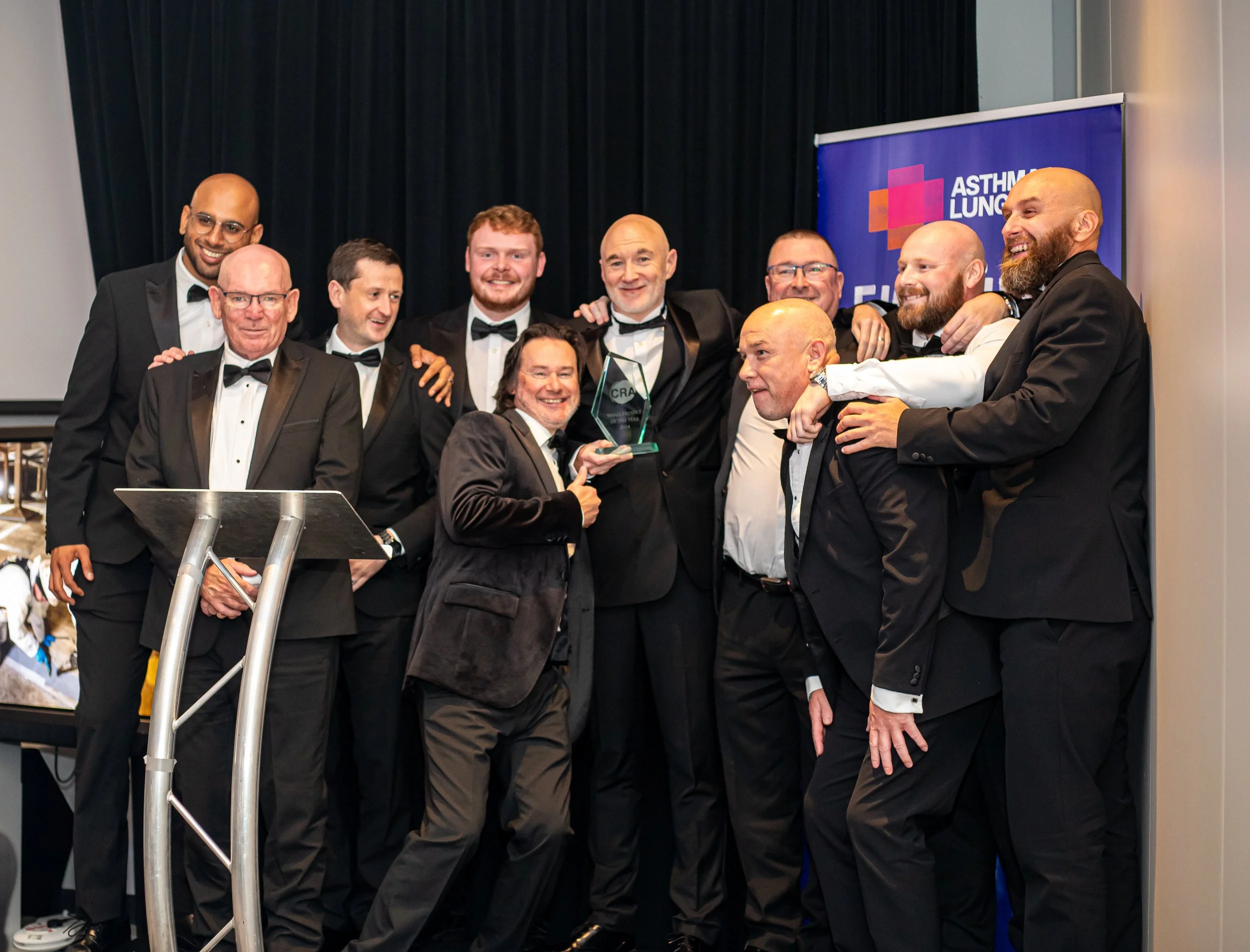 Group of nine men in tuxedos and bow ties, smiling and posing together at an awards event. They are holding a trophy in front of a blue banner with the text 'ASTHMA LUNG' and a colorful logo. The men are standing on a stage with a black curtain backg