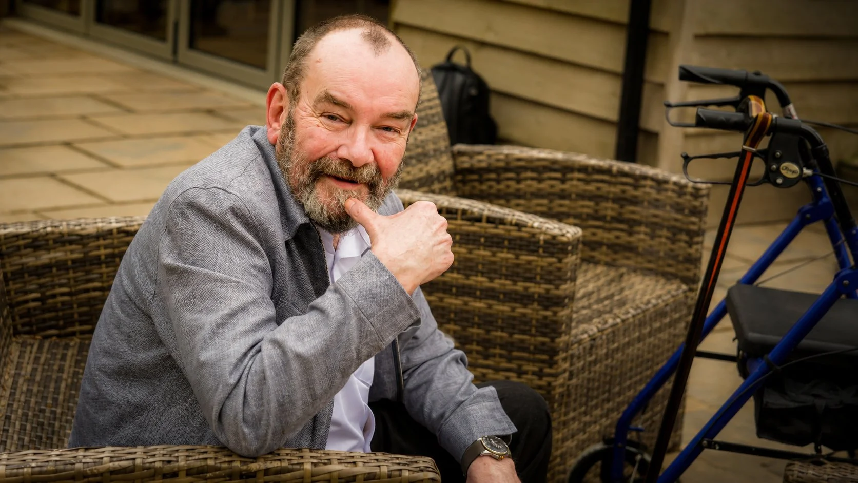 A smiling middle-aged man with a beard and receding hairline sitting outdoors on wicker furniture, giving a thumbs up, with a walker nearby.