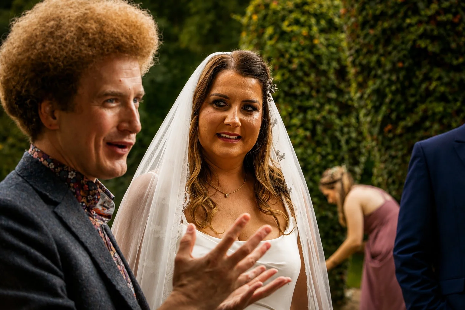 A bride with a veil and wedding dress talking to a man with reddish curly hair and a patterned shirt at an outdoor wedding ceremony.