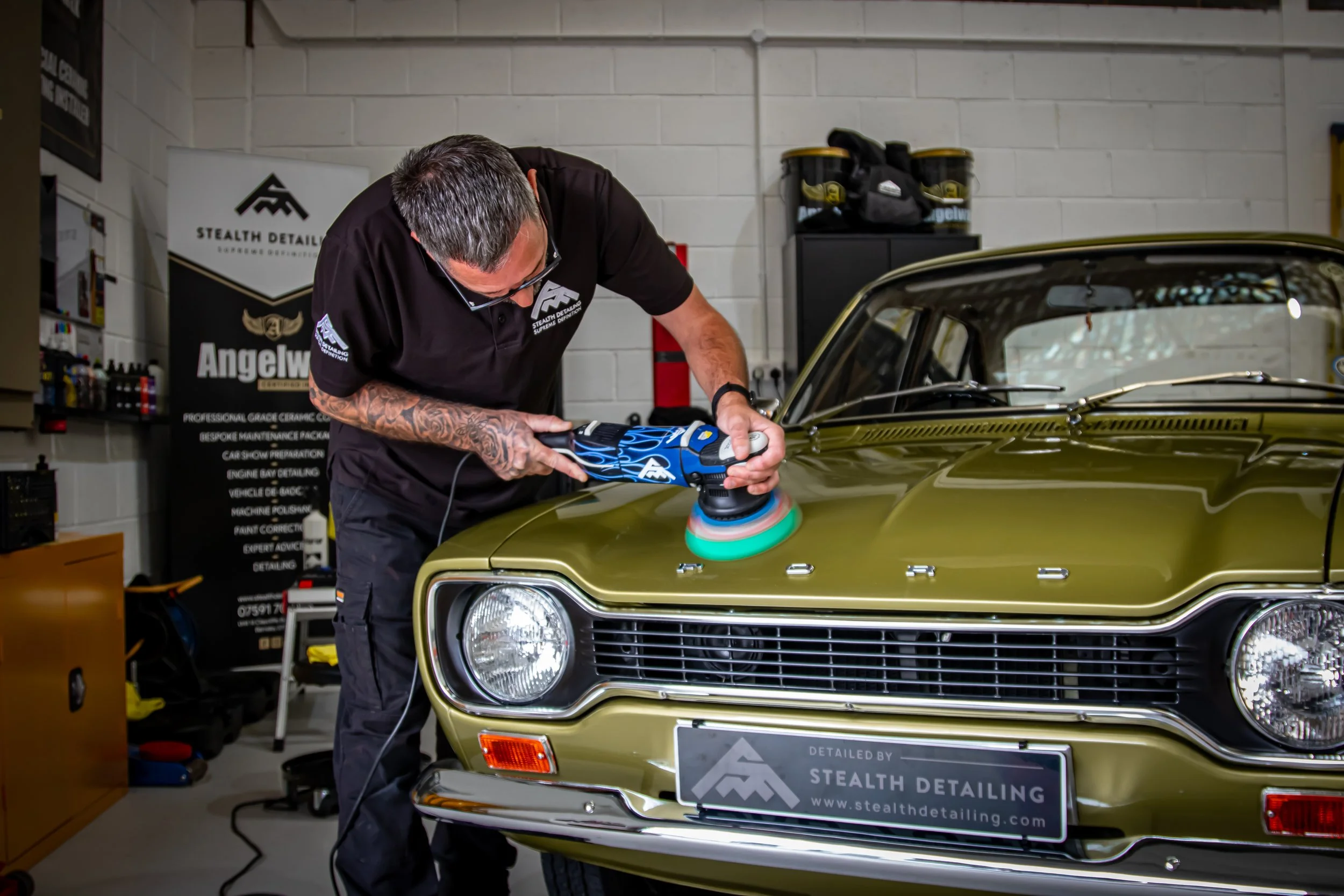 A man polishing a vintage gold-colored car with a handheld electric buffer in a garage. The garage has a sign that reads 'Stealth Detailing' and a black cabinet with car cleaning supplies.
