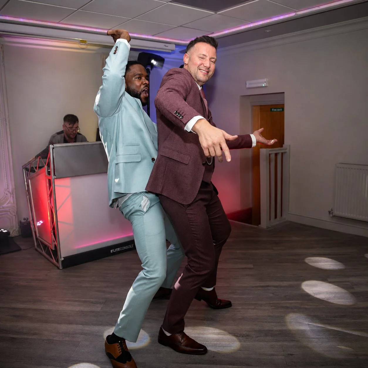 Two men dancing at a celebration or party, with a DJ in the background. One is wearing a light blue suit and the other a maroon suit, both enjoying and dancing energetically. The room has purple lighting and a wooden floor.