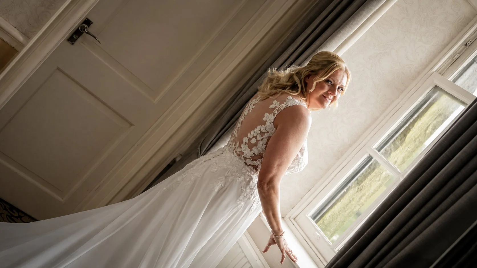 A bride in a wedding dress standing near a window, looking over her shoulder and smiling.