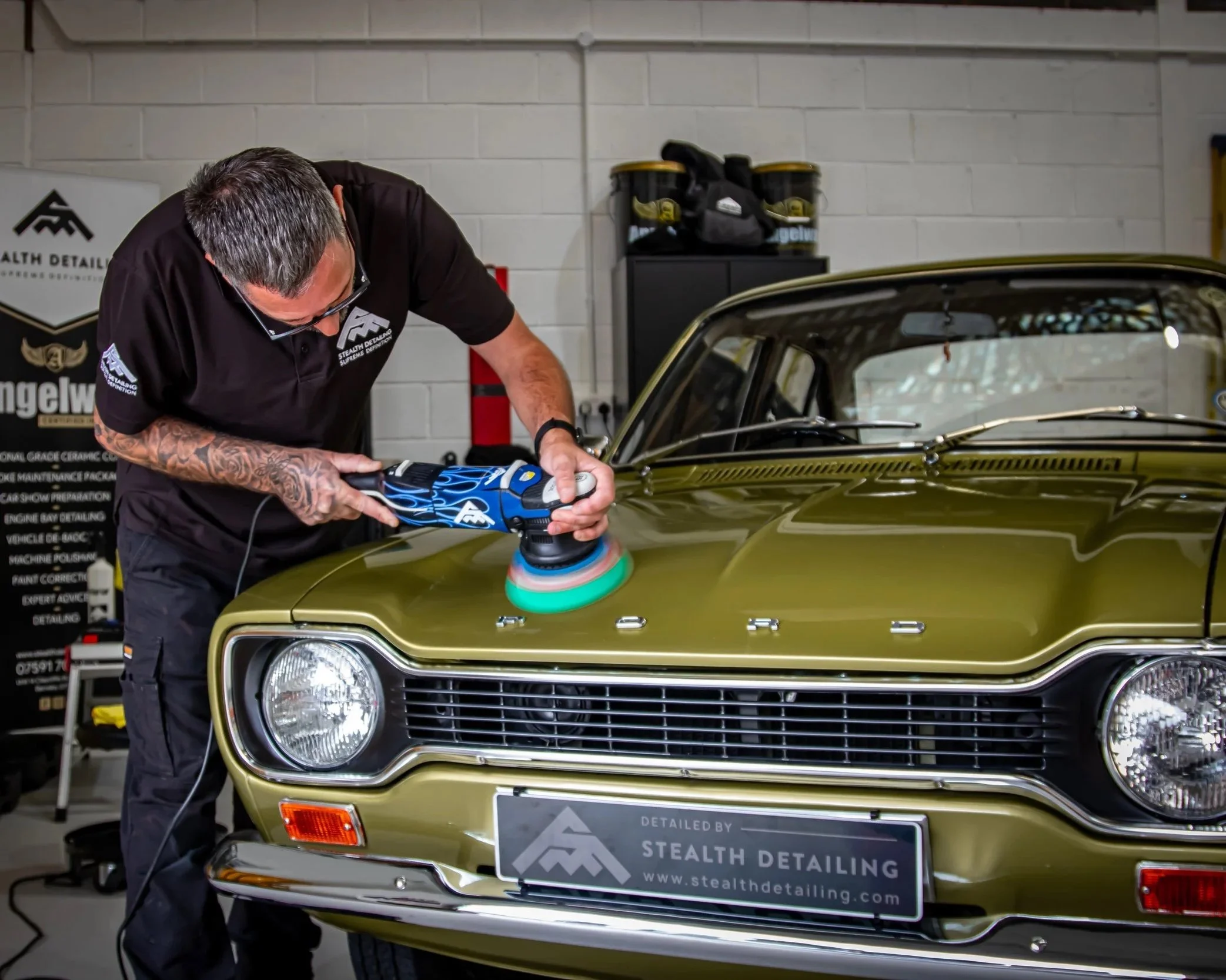 A man polishing a vintage gold-colored car with an electric buffer in a garage. The garage has a white brick wall, shelves with car detailing supplies, and a sign for Stealth Detailing.