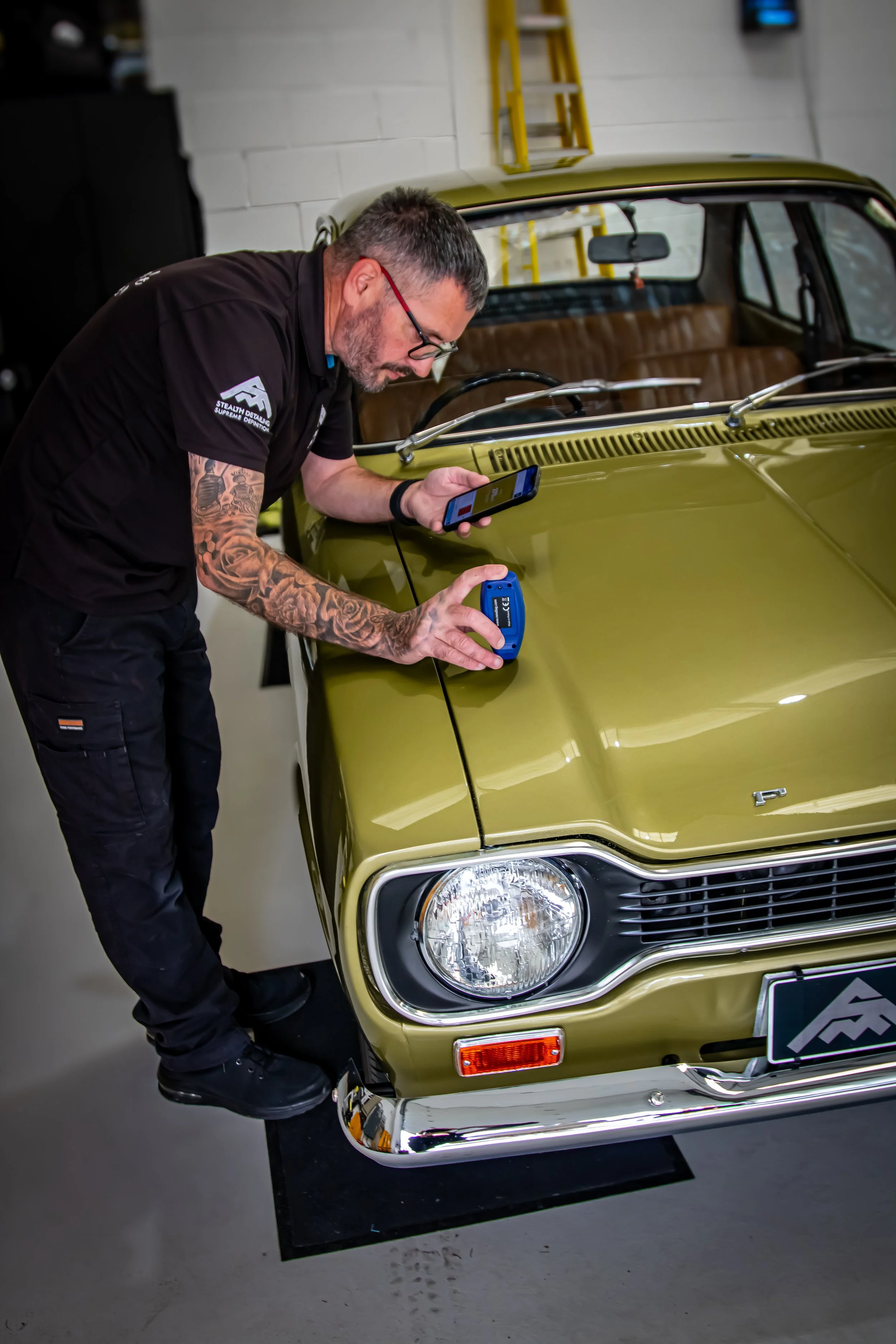 A man with tattoos on his arm and glasses is inspecting a vintage olive-green car using a handheld device and a smartphone, inside a garage.