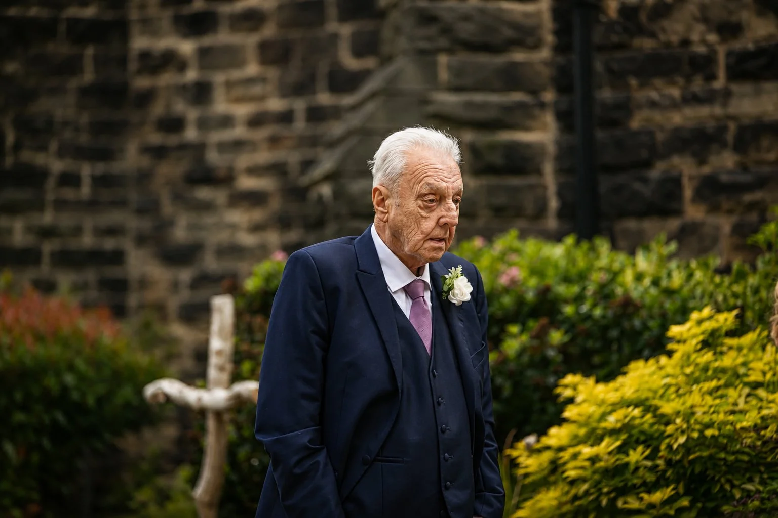 An elderly man with white hair wearing a navy suit, light purple tie, and a white boutonniere stands outside against a stone wall, surrounded by bushes and greenery.
