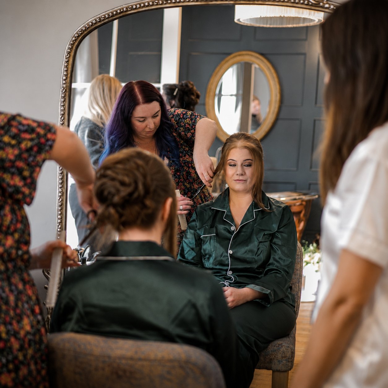 A woman with auburn hair is getting her hair styled by a hairstylist in a salon, with several people gathered around.