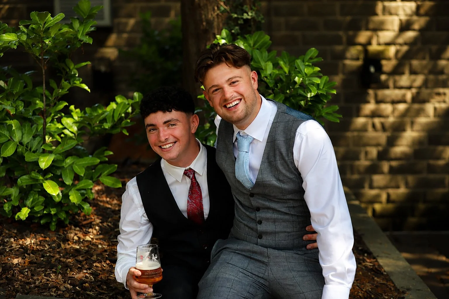 Two men in formal attire smiling outdoors, one holding a glass of beer, surrounded by green bushes and a brick wall in the background.