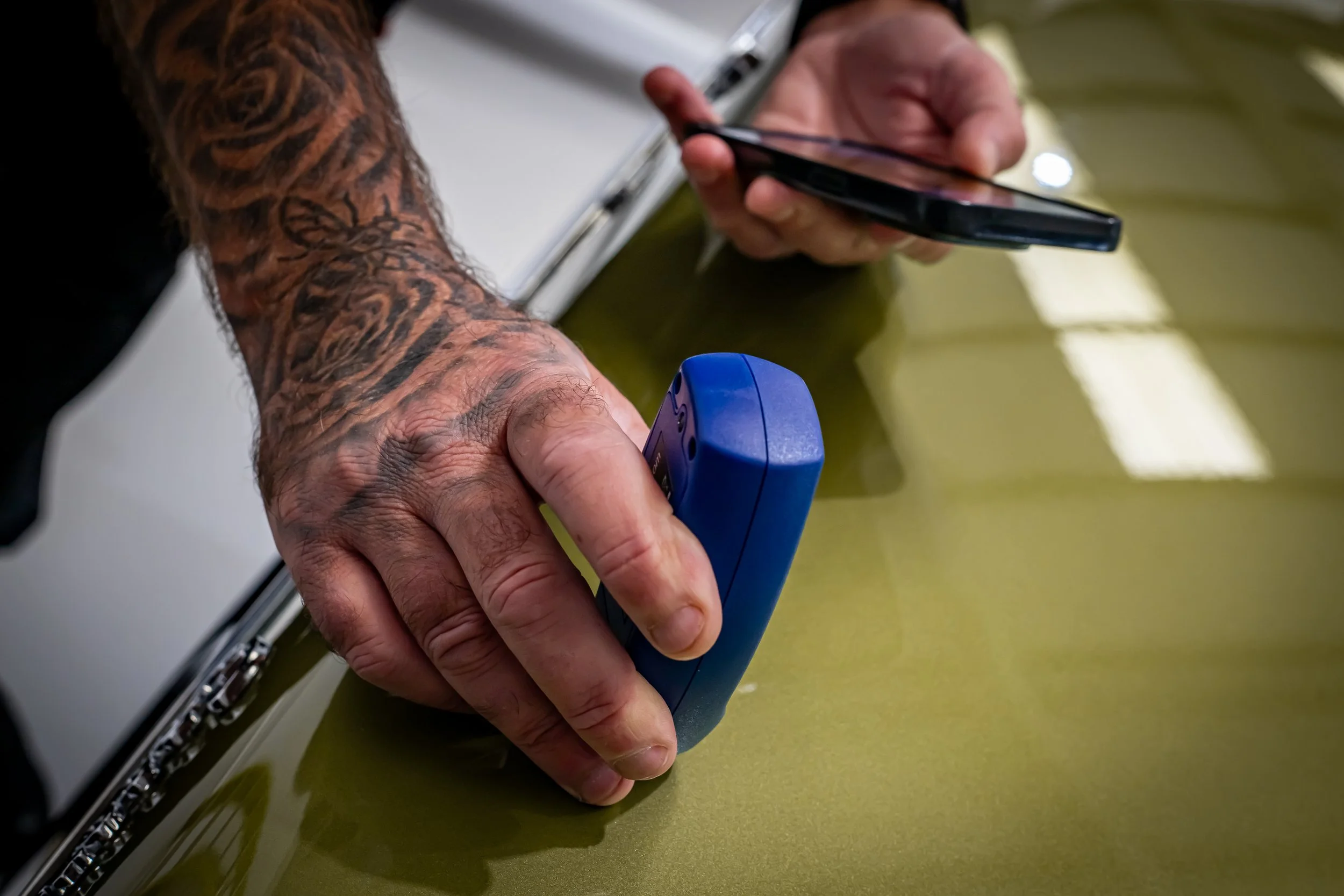 A person's tattooed arm holding a blue device, while another person’s hand holds a smartphone, with a white sheet of paper partially visible in the background, on a green surface.