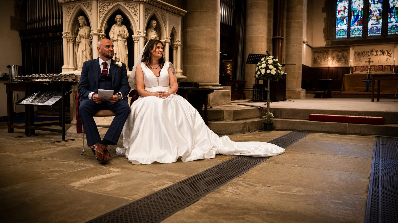 Bride and groom seated during a wedding ceremony in a church with stained glass windows and gothic architectural details.