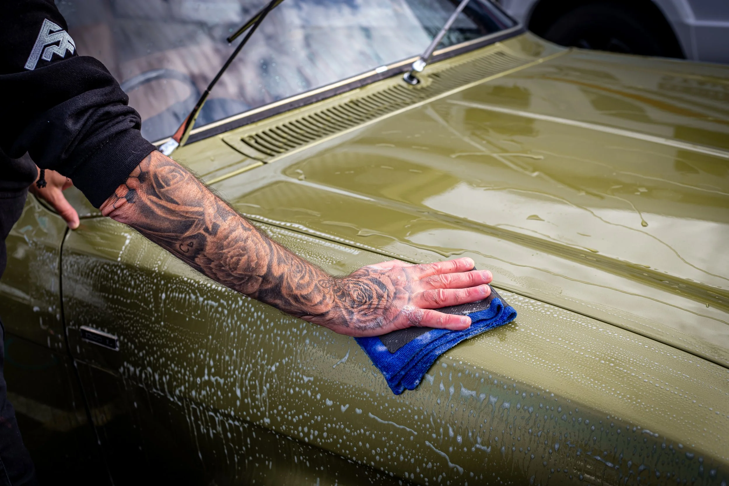 Person with tattooed arm washing a green car with a blue cloth, soap suds and water visible.