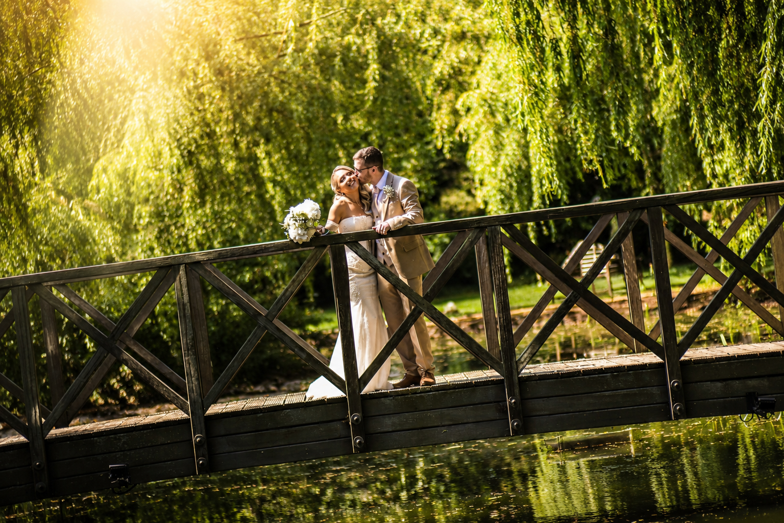 A newlywed couple standing on a wooden bridge over a pond, embracing and sharing a kiss. The bride holds a bouquet of white flowers. The scene is surrounded by lush green trees and illuminated by sunlight.
