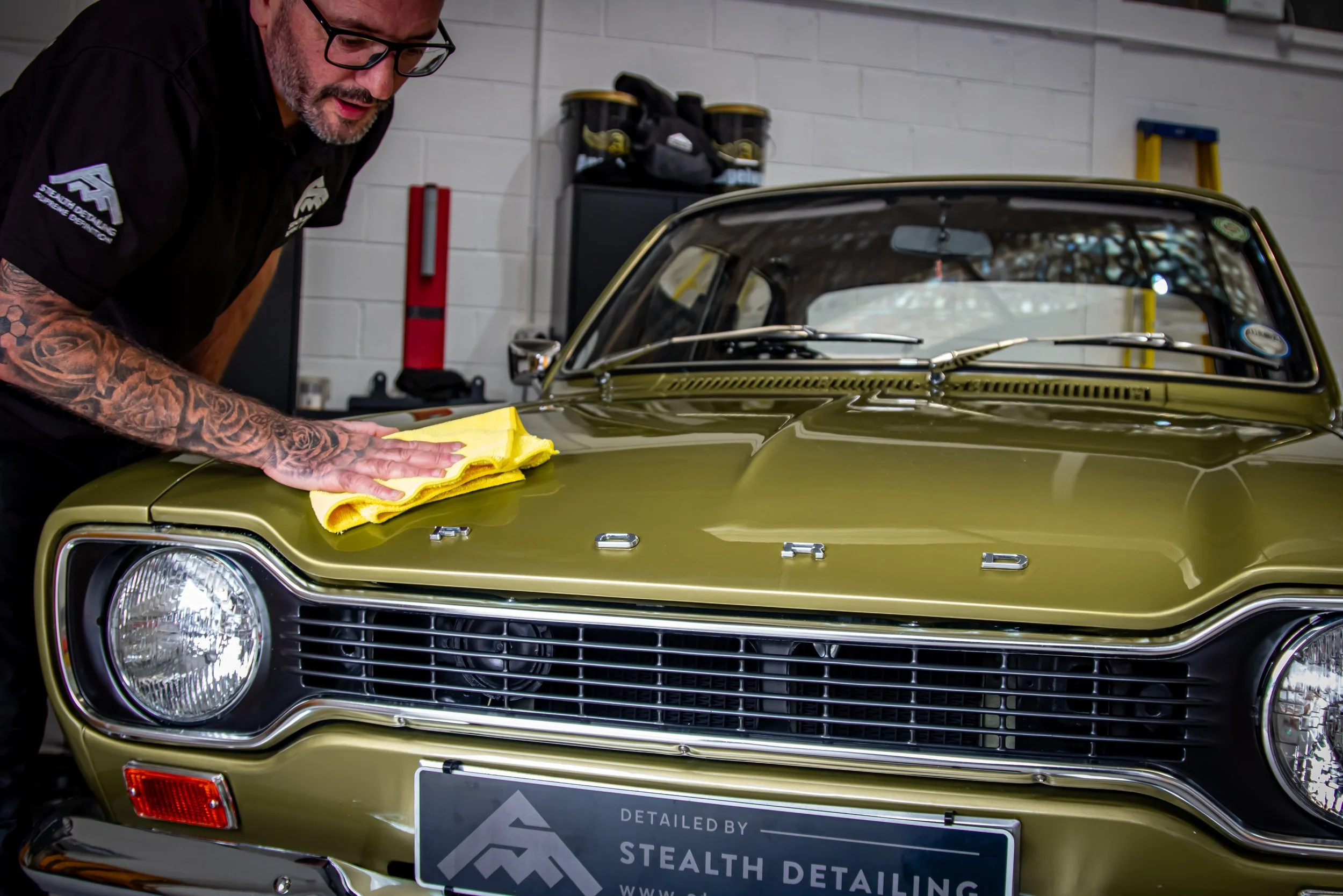 A man with glasses and tattoos on his arm is cleaning or detailing an olive-green vintage Ford car inside a garage. He is holding a yellow cloth and leaning over the hood of the vehicle. The front grille and headlight of the car are visible, and the 