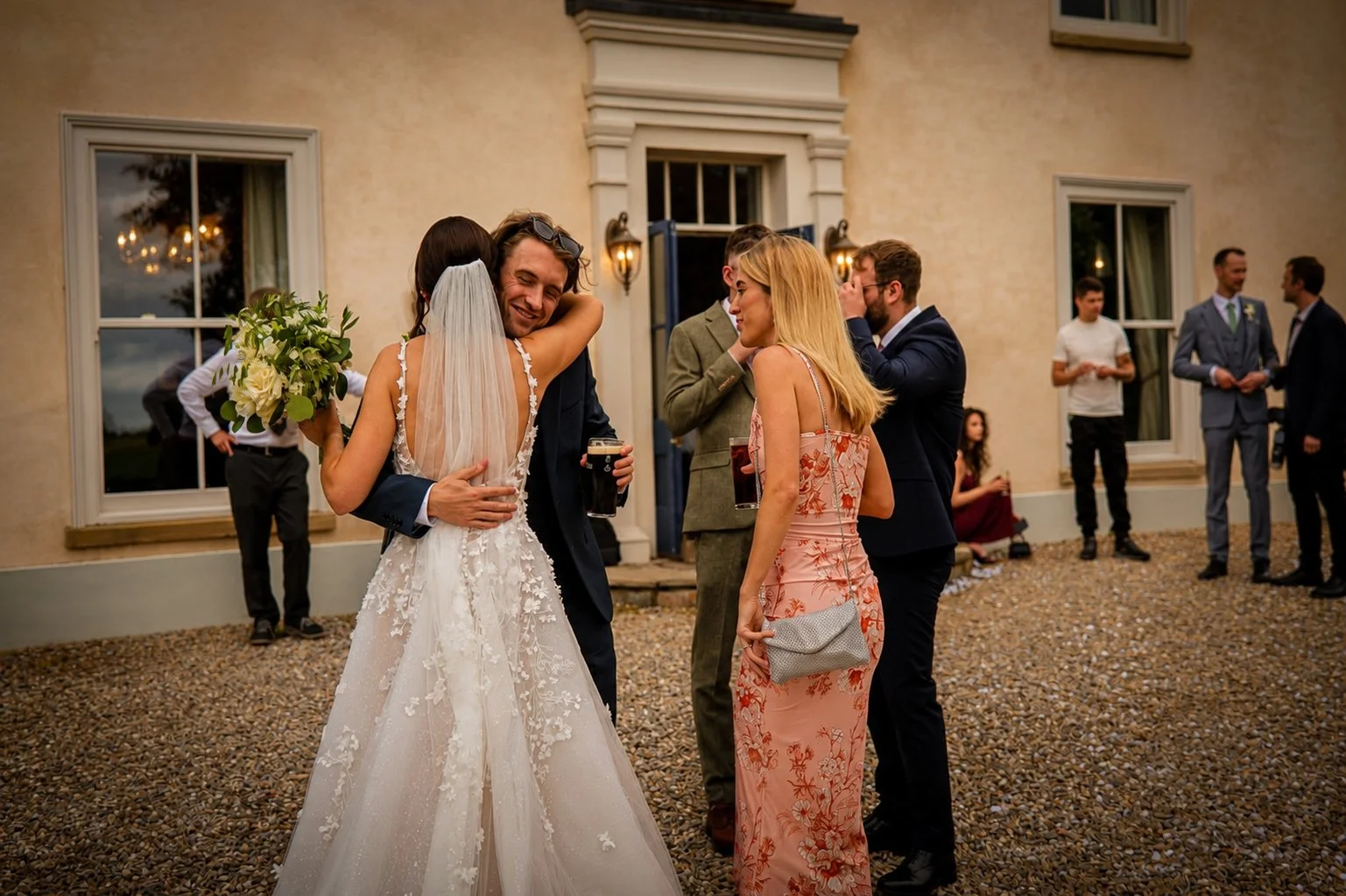 A bride in a white wedding gown hugging a man in a black suit holding a drink, surrounded by guests at an outdoor wedding reception with a beige building in the background.
