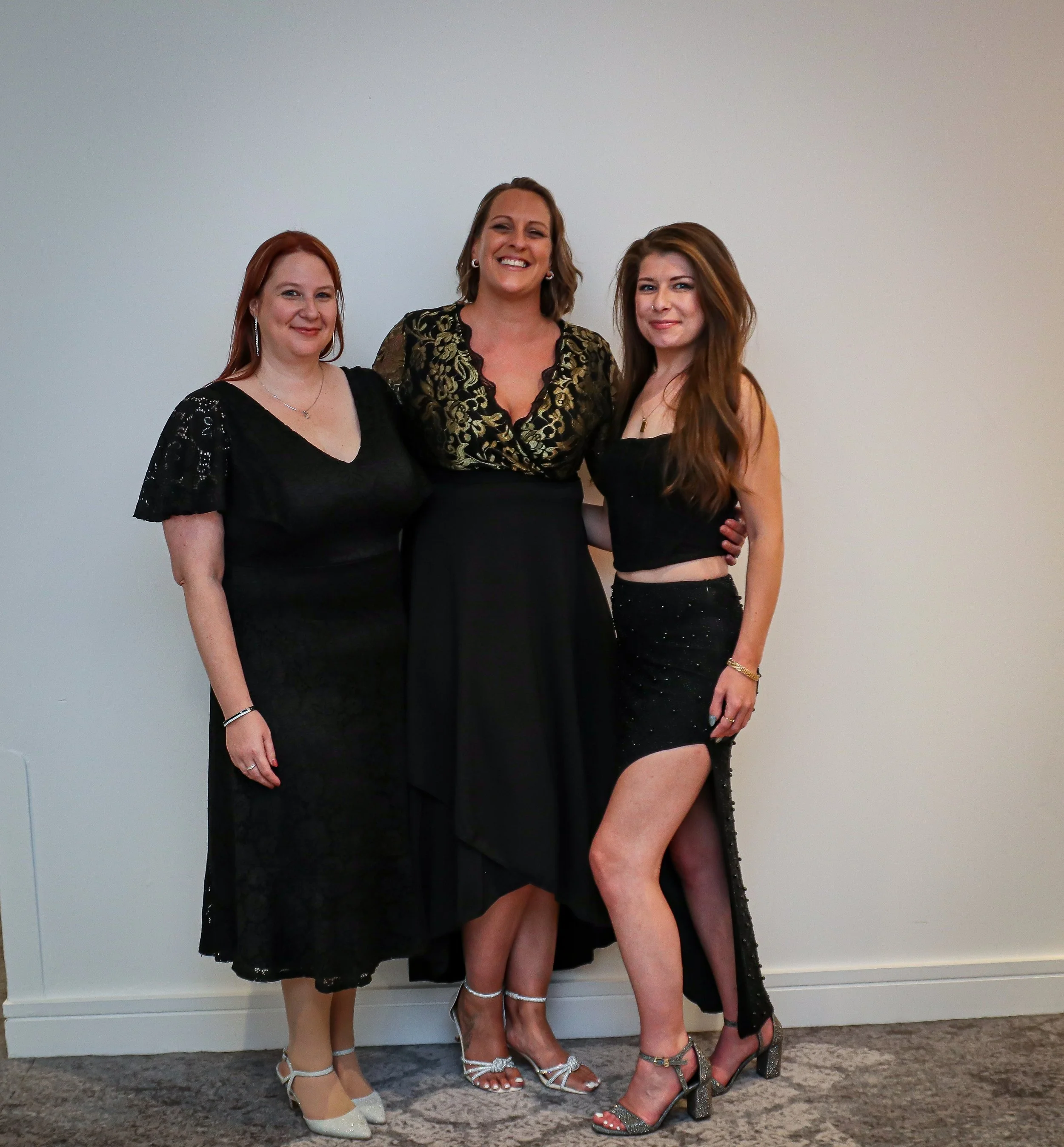 Three women dressed in formal black attire standing together indoors against a plain white wall, smiling for the camera.