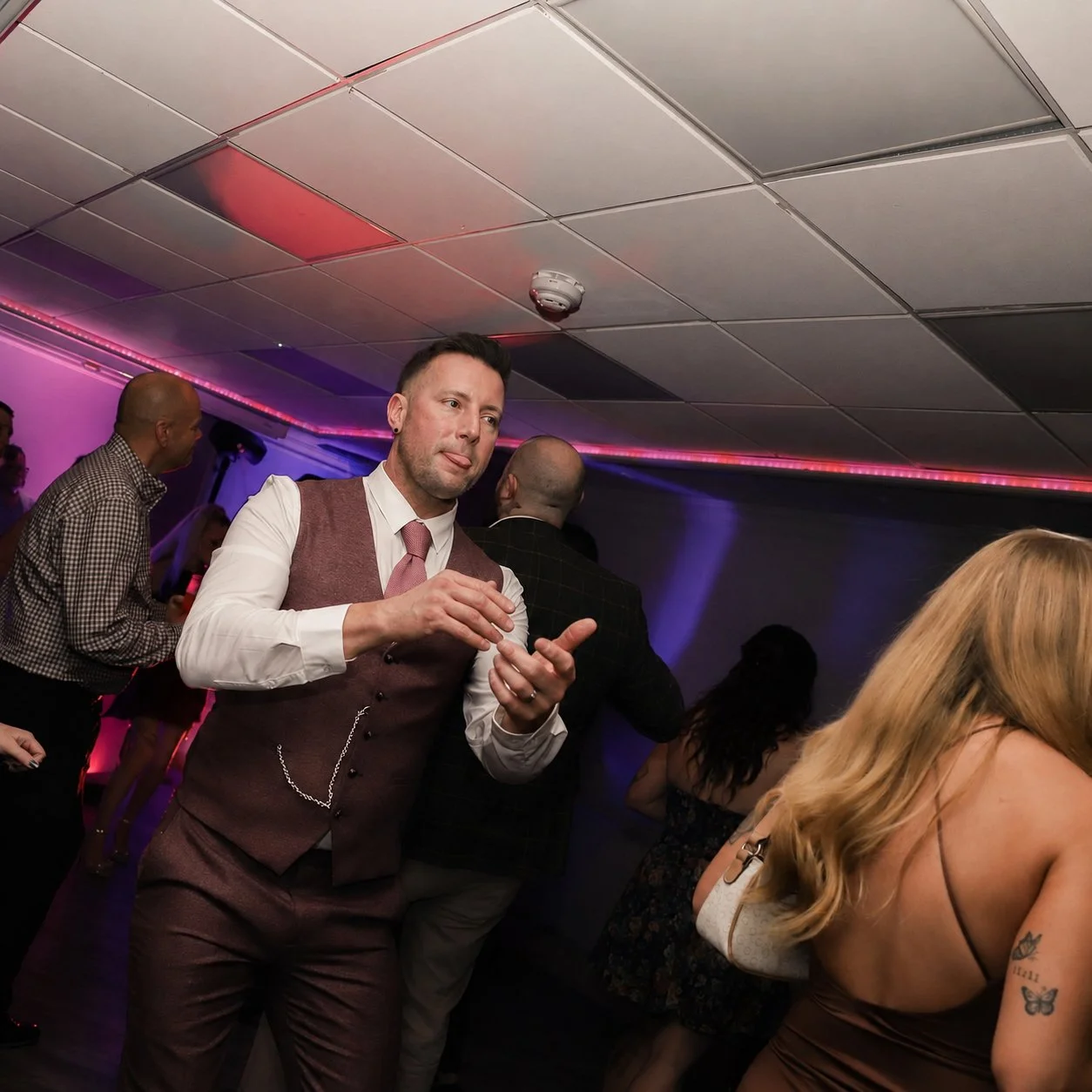 People dancing and socializing at a party with pink and purple lighting, including a man in a brown suit with a pink tie in the foreground.