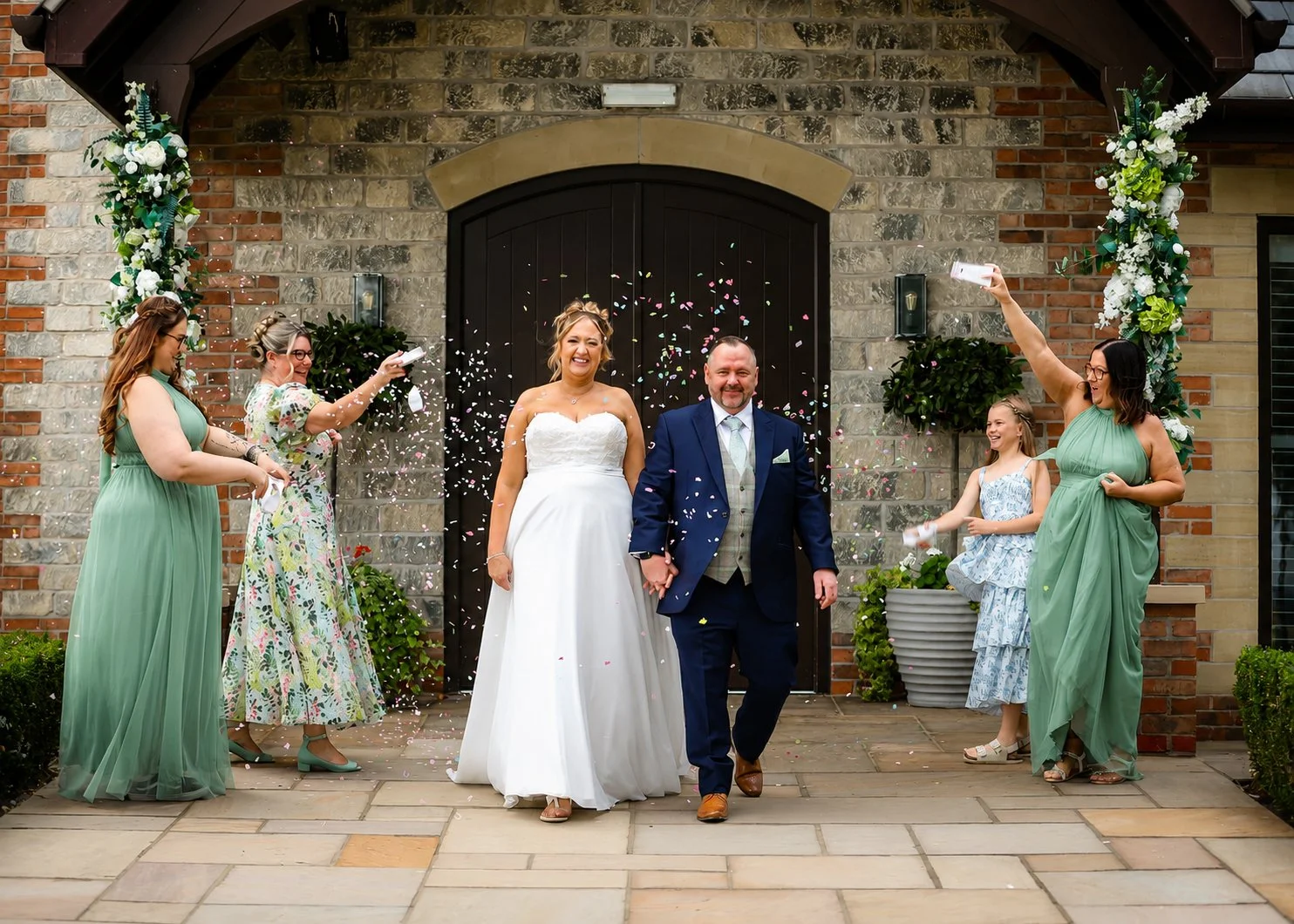 A wedding celebration with the bride and groom walking hand in hand outside a brick building, surrounded by friends throwing confetti. The bride is wearing a white strapless dress, and the groom is dressed in a dark suit with a tie and vest. The frie