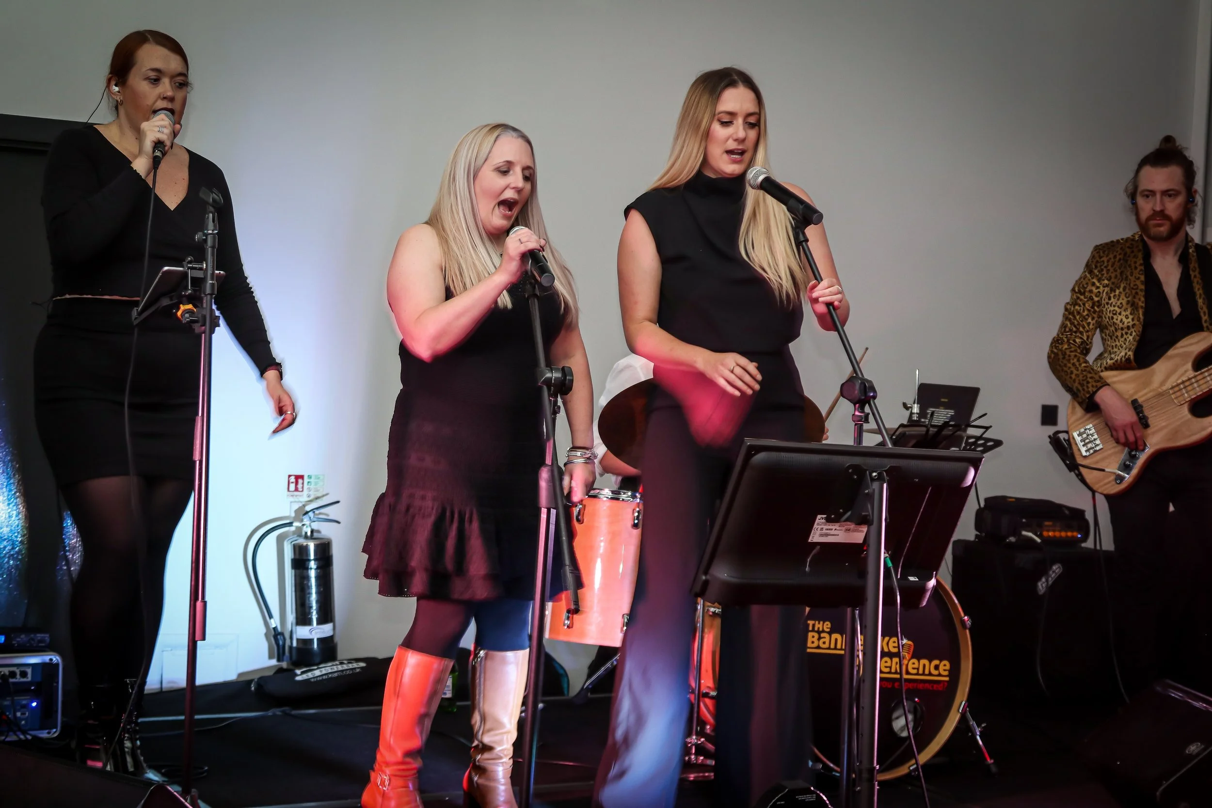 Four musicians performing on stage with microphones, a drum, and a bass guitar, against a neutral background.