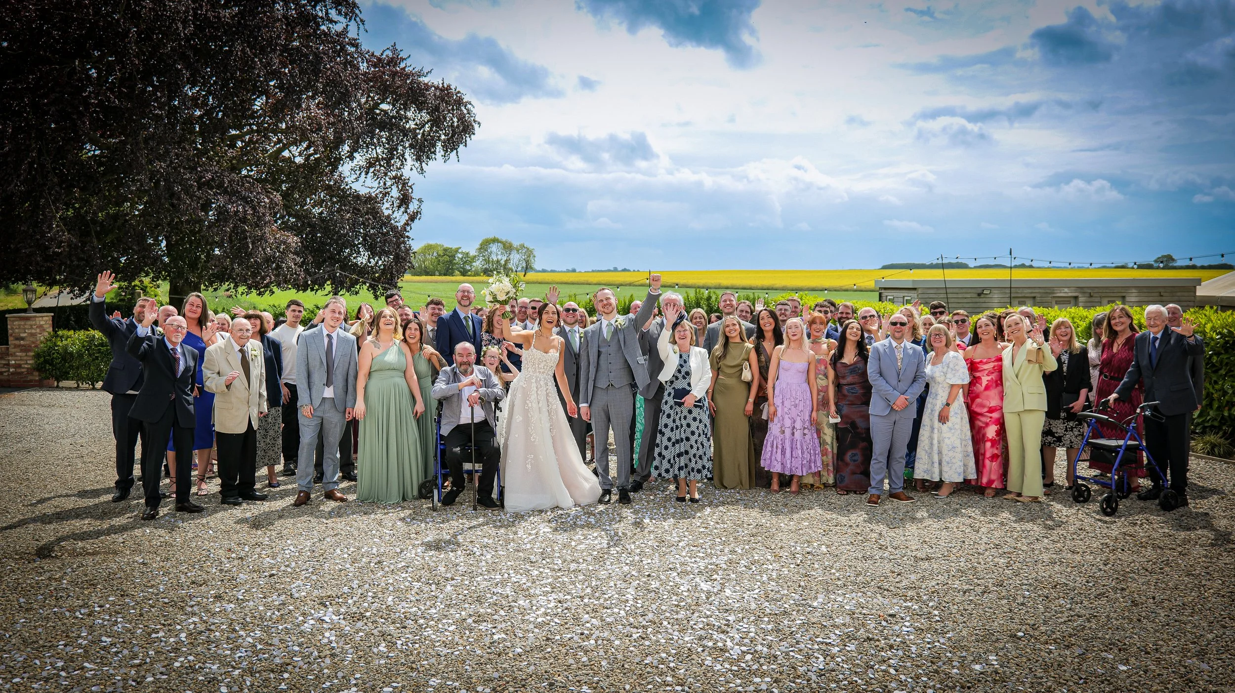 A large group of people, including a bride and groom, gathered outdoors for a wedding celebration under a large tree with a rural landscape background, some raising their hands and smiling.
