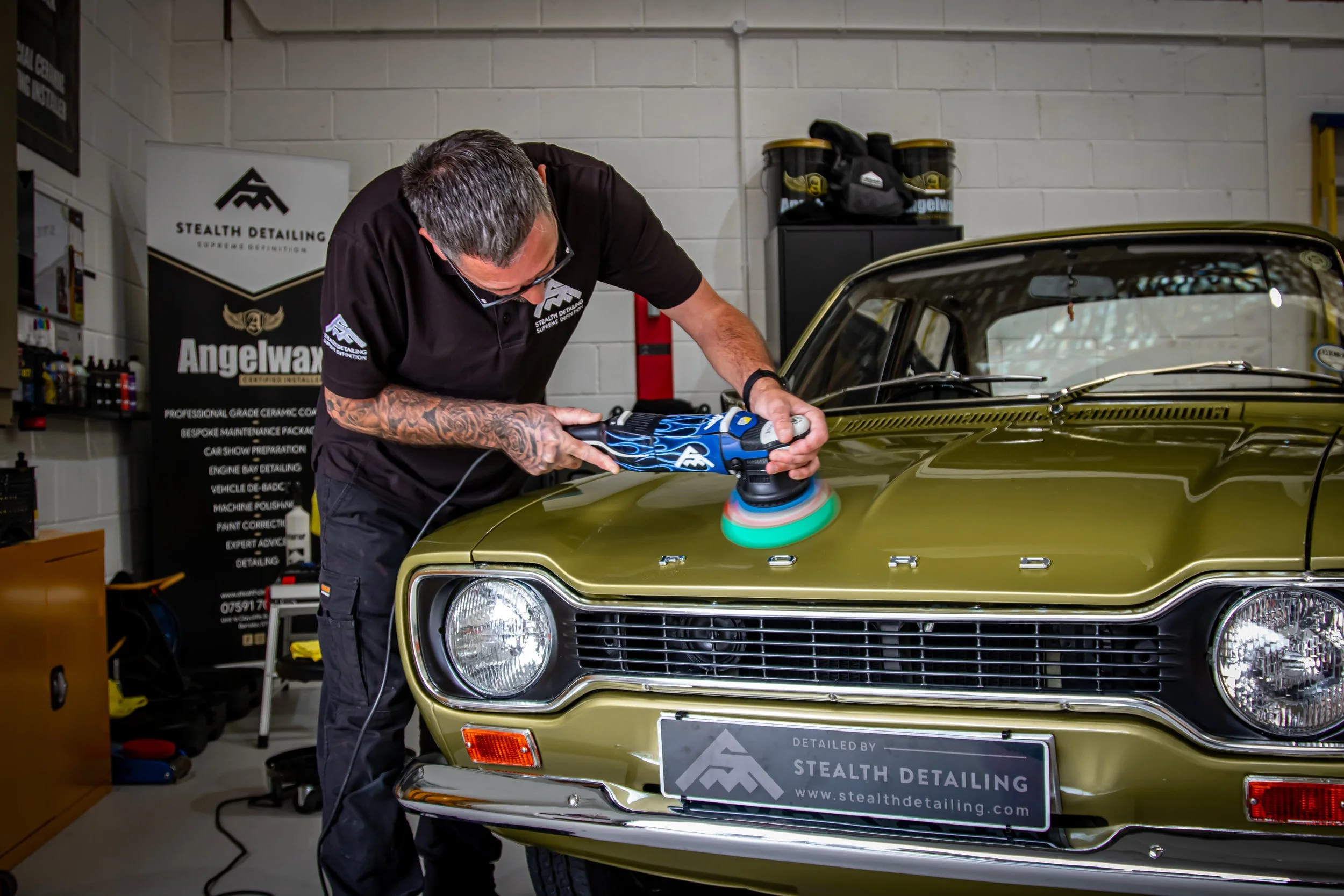 A man is polishing a vintage gold car with a polishing machine inside a garage. He is wearing glasses and a black polo shirt with company logos, focusing on the car's hood.