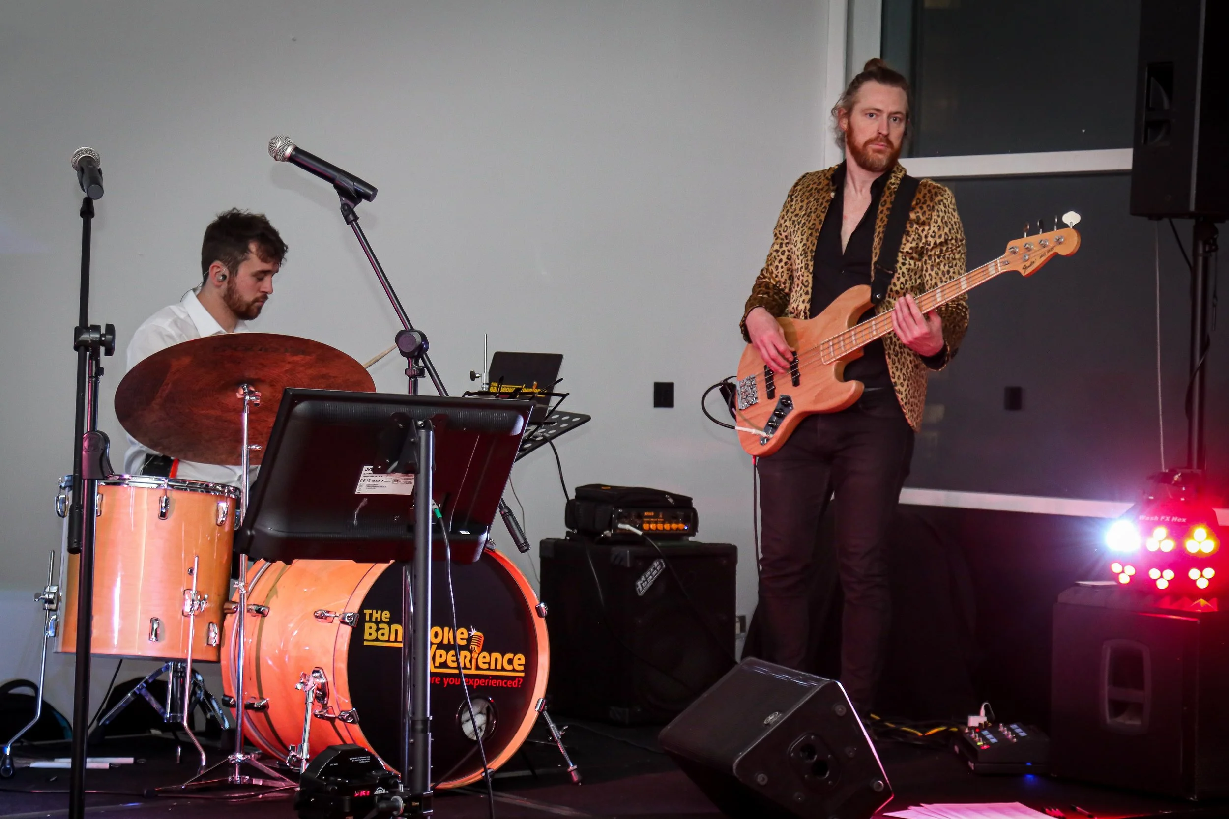 Two musicians performing on stage: one playing the drums and the other playing an electric bass guitar. The drummer is sitting behind a drum set with a bass drum that has a logo and words on it. The bassist is standing, wearing a leopard print jacket