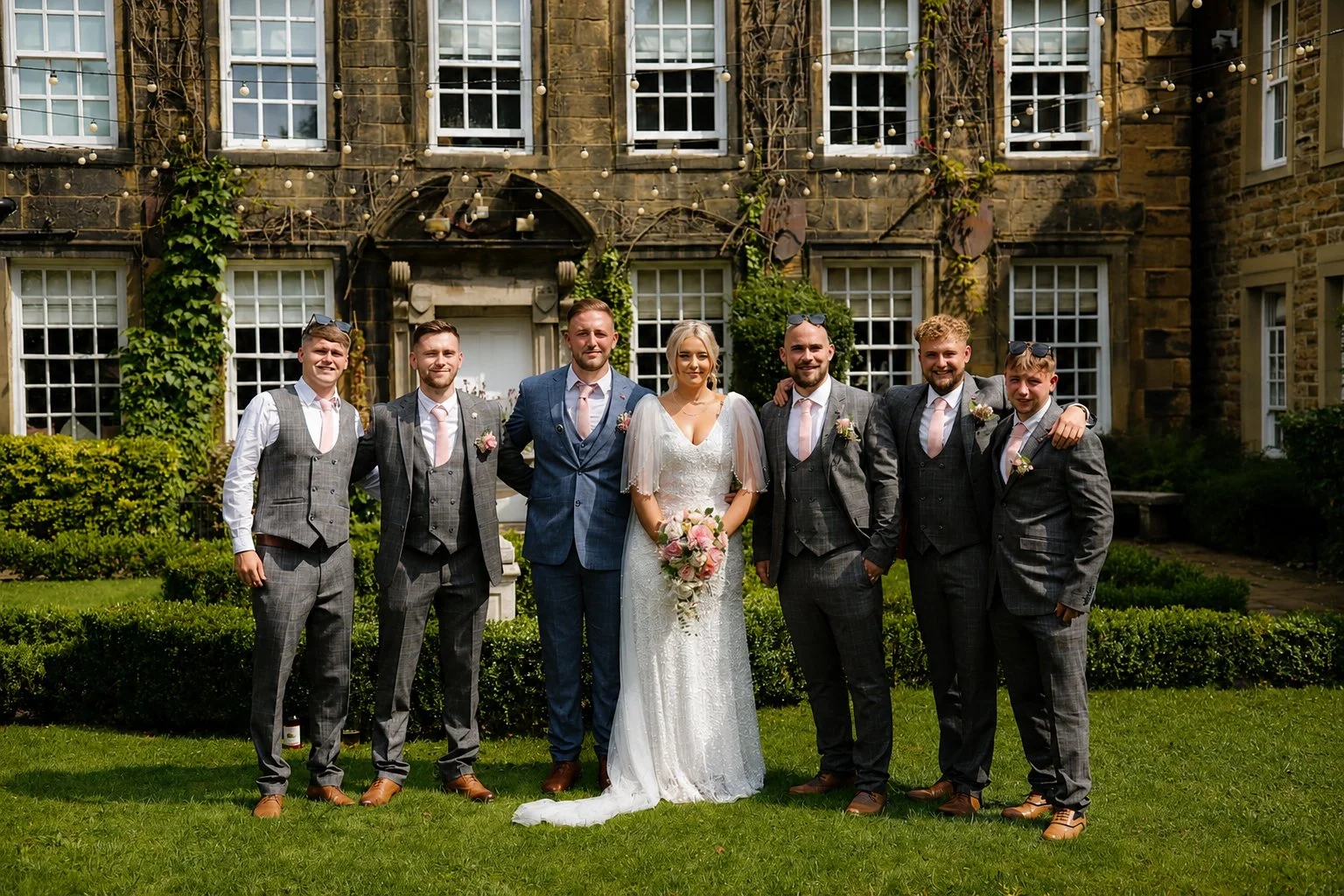 Group of people in wedding attire standing on a lawn in front of a historic stone building with multiple windows and green bushes.