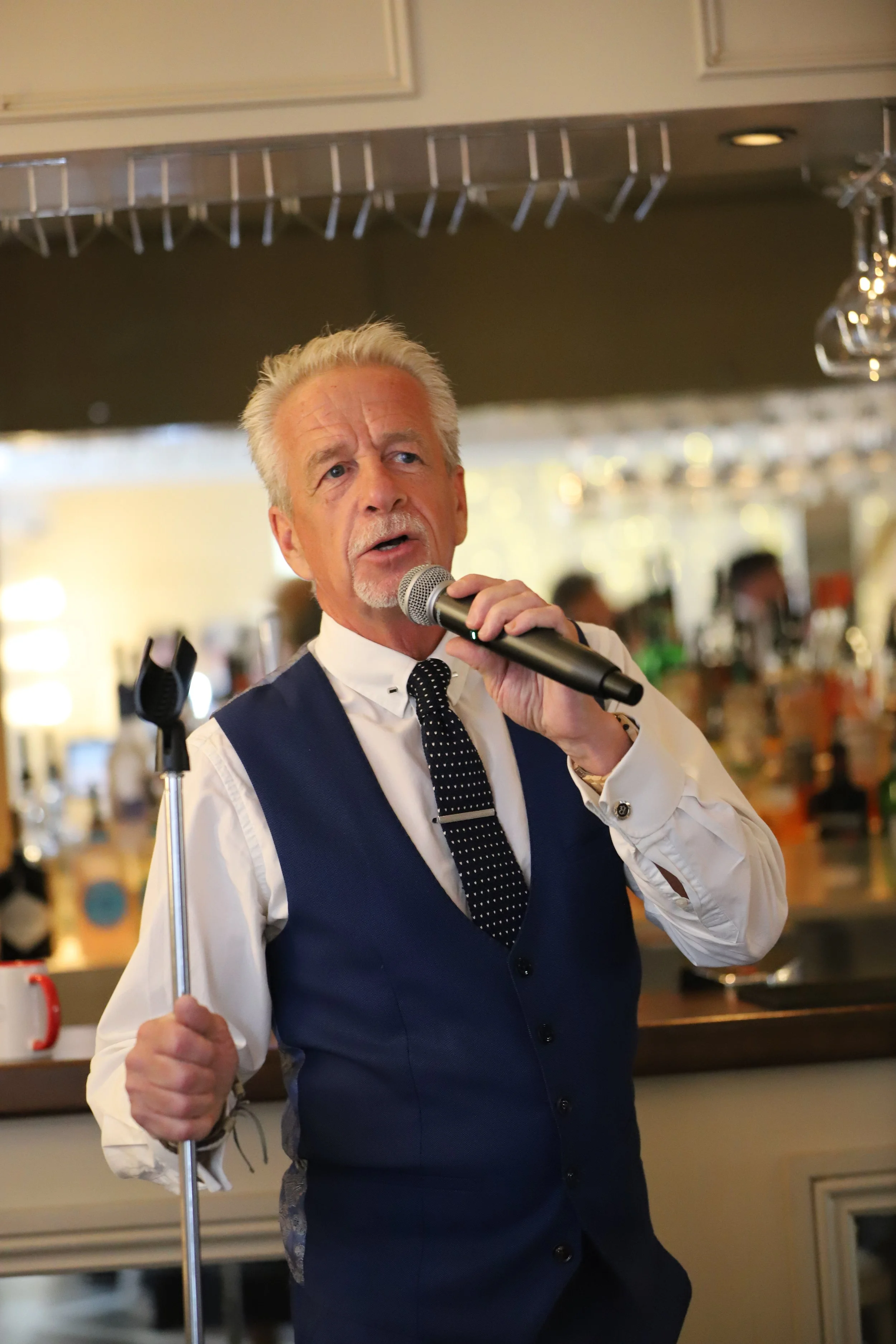 An older man with gray hair and a mustache wearing a white shirt, navy vest, and polka dot tie, holding a microphone and speaking.