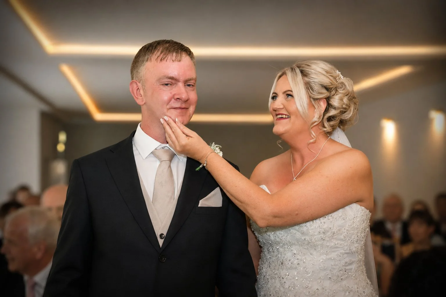 A bride with an elegant updo hairstyle and wedding dress touching the face of a groom in a suit during a wedding ceremony.