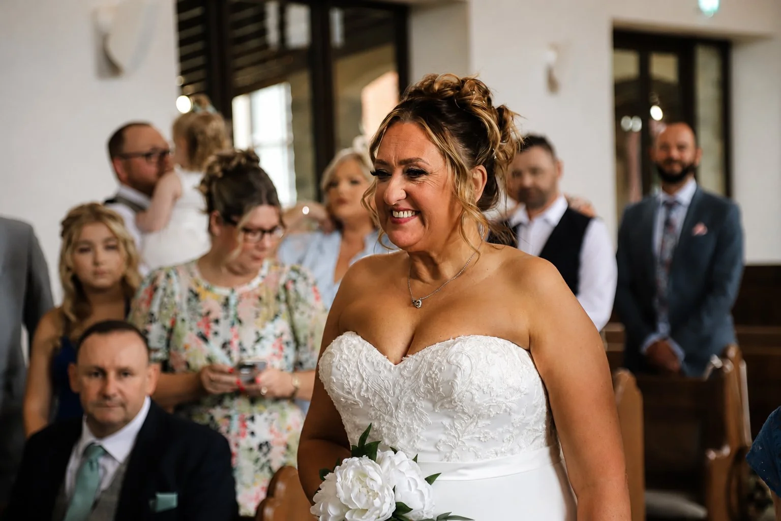 A bride on her wedding day, smiling and holding a bouquet, in a church filled with family and friends.
