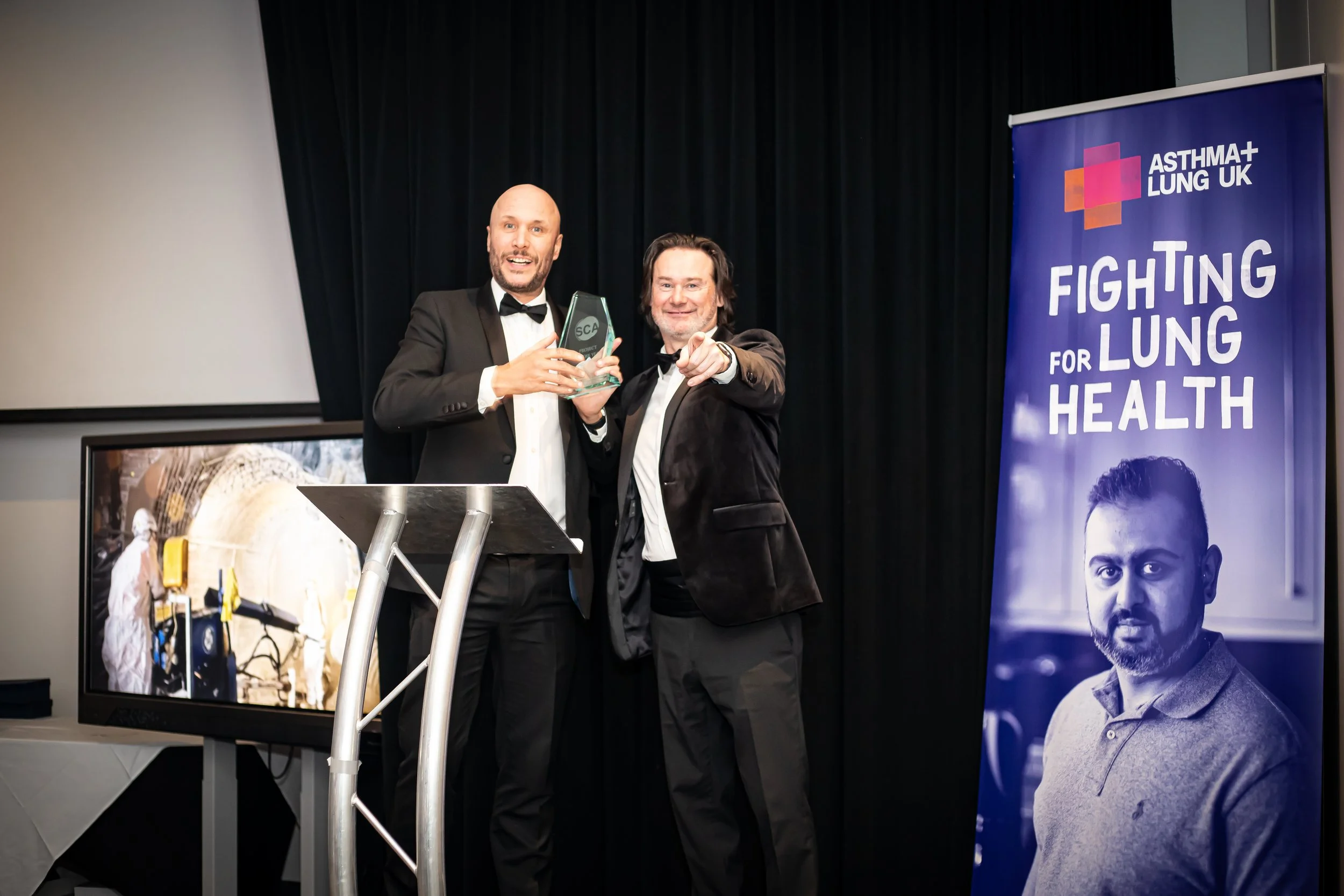 Two men in tuxedos at an award ceremony, one holding a glass award, smiling at the camera, with a banner reading 'Fighting for Lung Health' and a large screen displaying an image of a tunnel construction site.