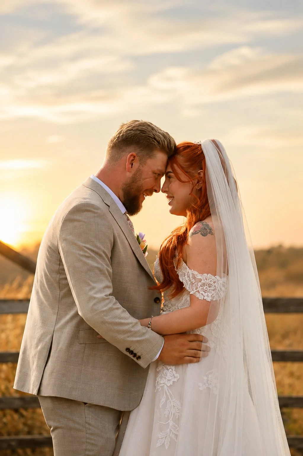 A newlywed couple, a man in a beige suit and a woman with red hair and a white wedding dress, are smiling and touching foreheads during sunset outdoors.