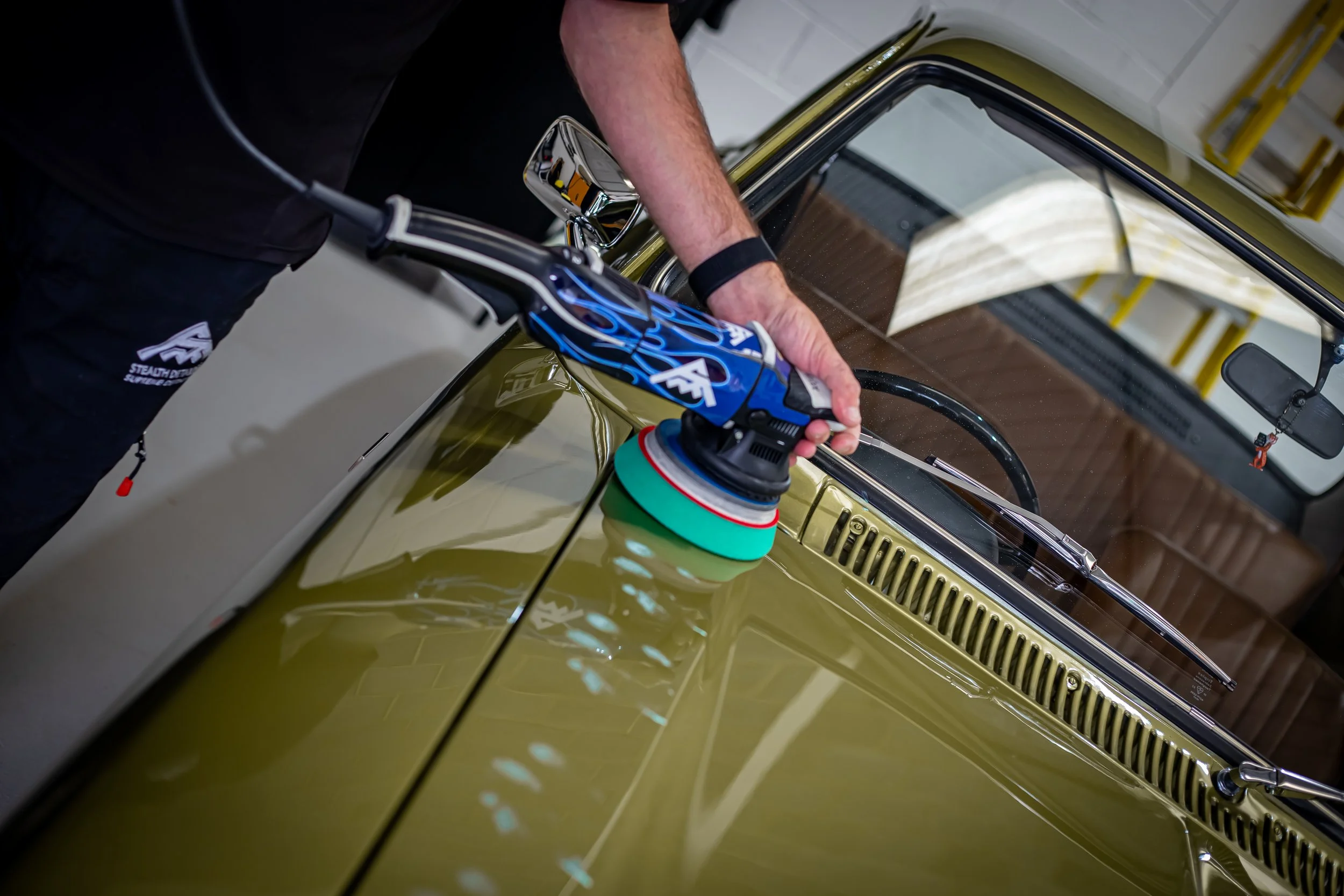 A person polishing a green vintage car with a power buffer in a garage.