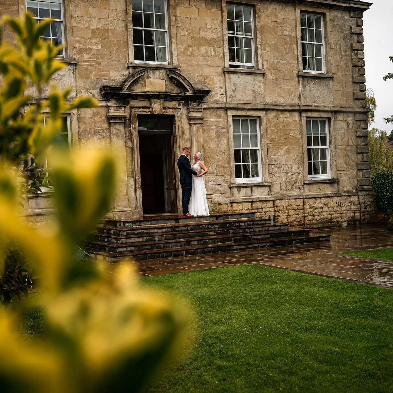 A bride and groom standing on outdoor stairs in front of a stone building, holding hands and facing each other during a wedding photoshoot. The building has six tall windows and a large front door.