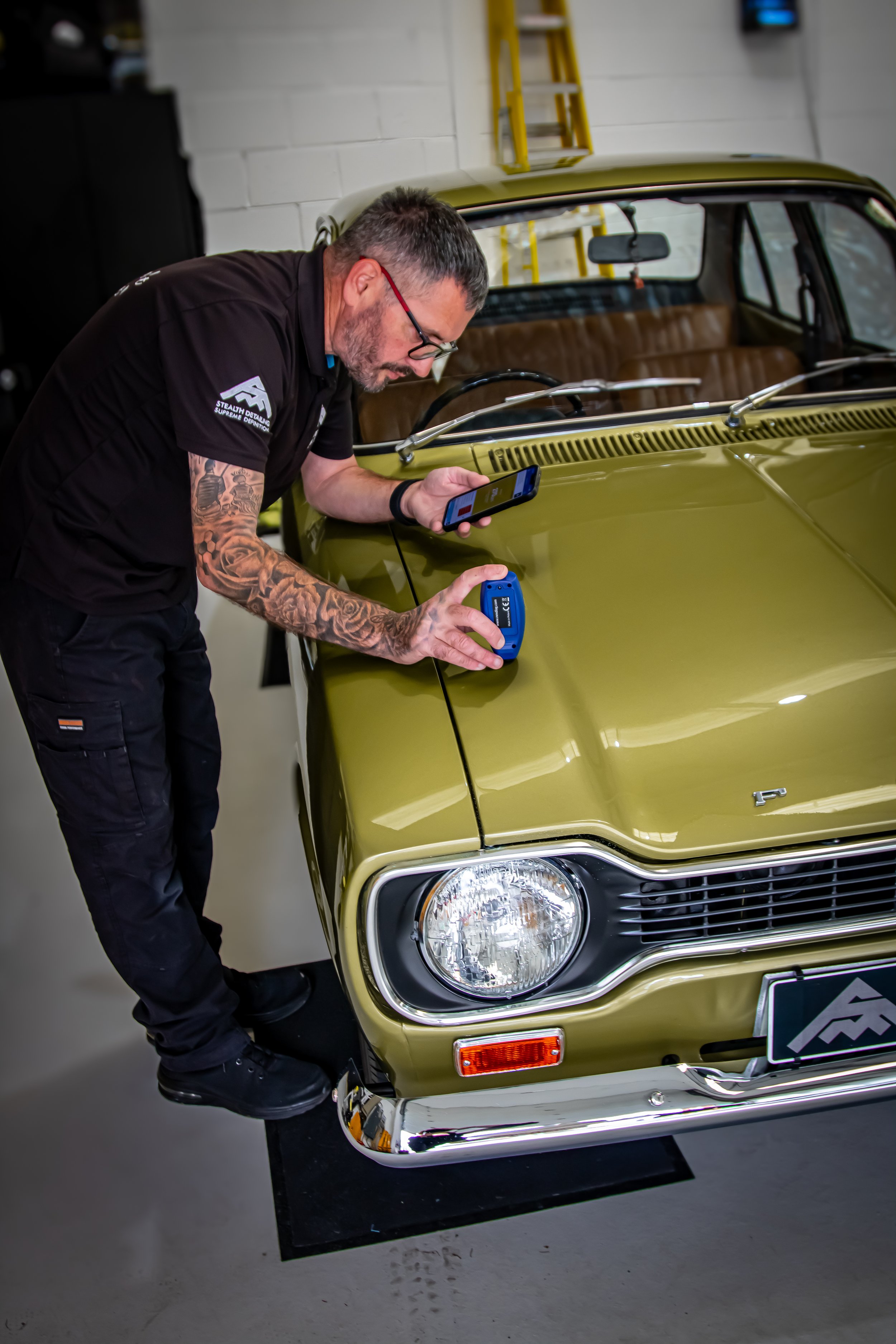A man with tattoos on his left arm and glasses is working on a vintage gold-colored car inside a garage. He is holding a diagnostic scanner in his left hand and using a smartphone in his right hand, looking down at the car's hood.