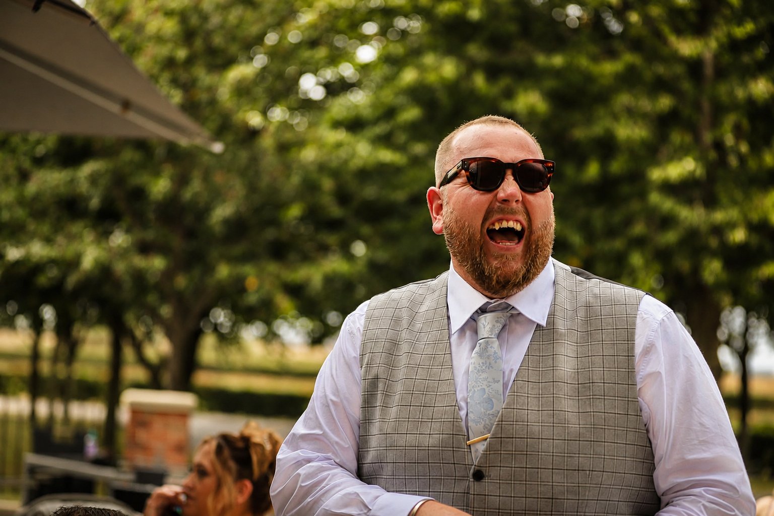 A man with a beard wearing sunglasses, a white shirt, a tie, and a gray vest, with an open mouth, speaking or shouting outdoors during daytime with trees in the background.