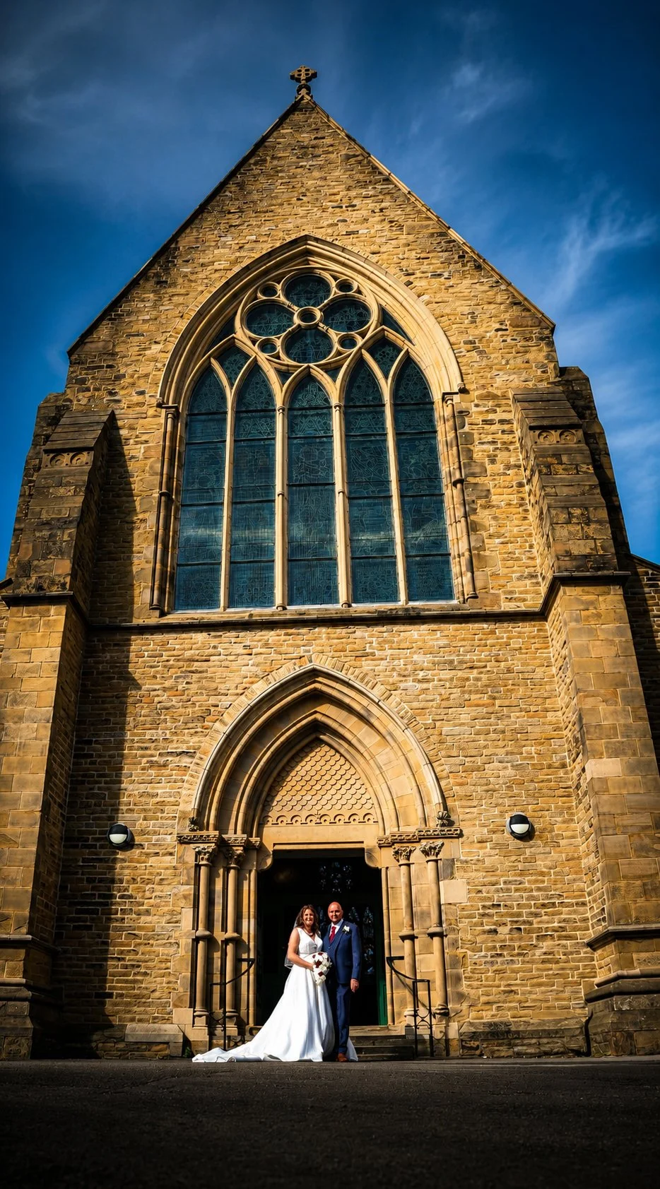 A bride and groom standing in front of a large stone church with stained glass windows, during sunset.