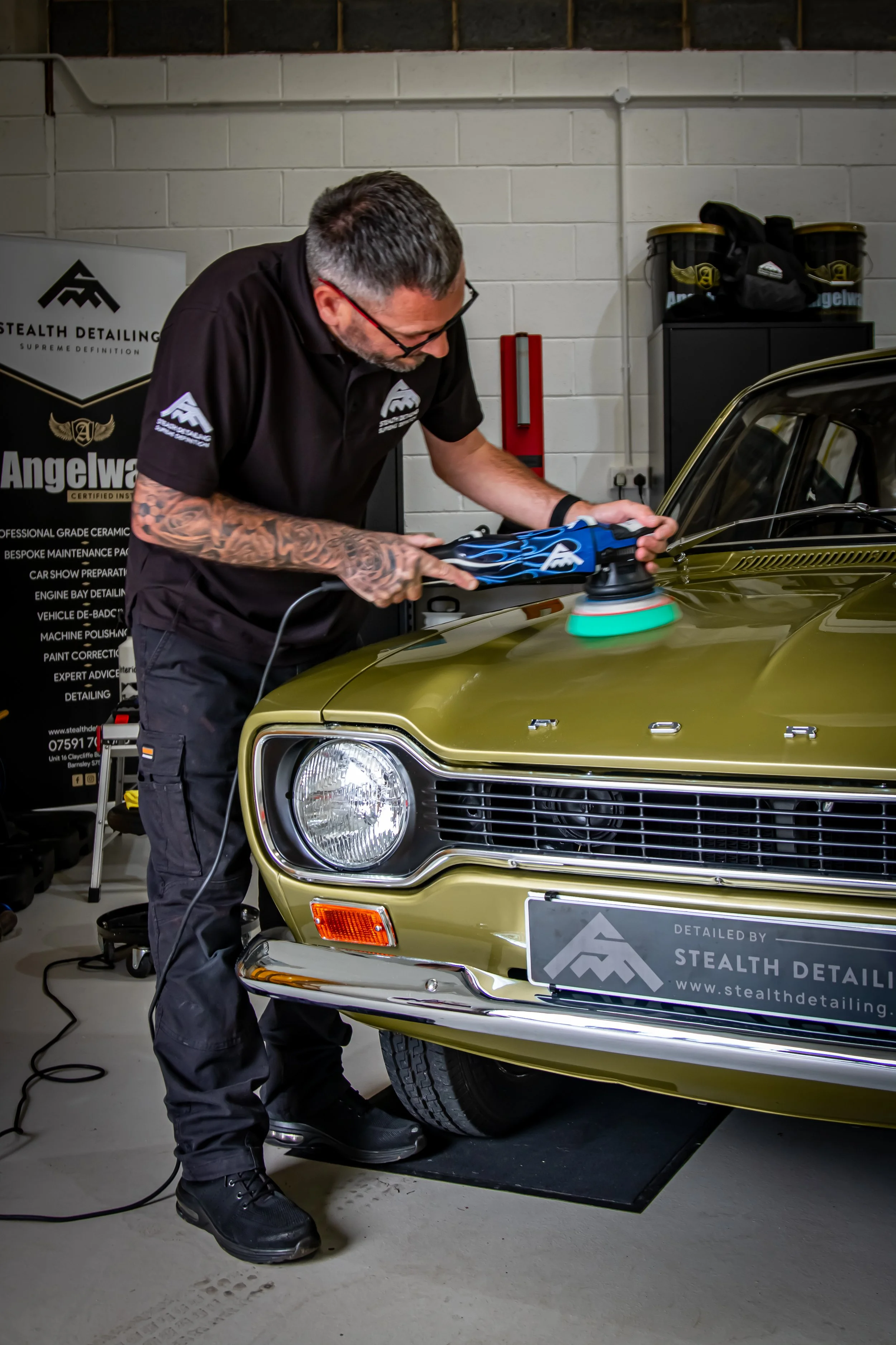 A man with tattoos, glasses, and dark clothing is polishing a vintage gold Ford Mustang using a blue electric polisher with a foam pad. He is inside a garage with white brick walls, a black storage cabinet, and a Stealth Detailing banner in the background.