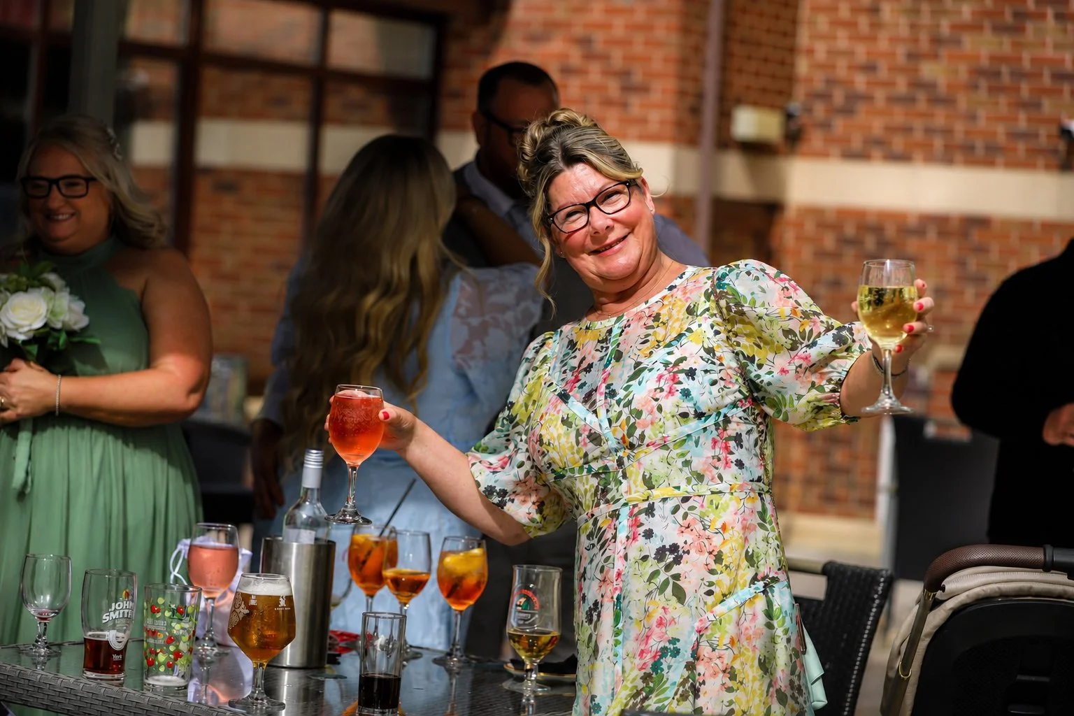 A woman wearing glasses and a floral dress, smiling and holding a glass of white wine and a glass of rosé, standing at a table with various drinks at a social gathering.