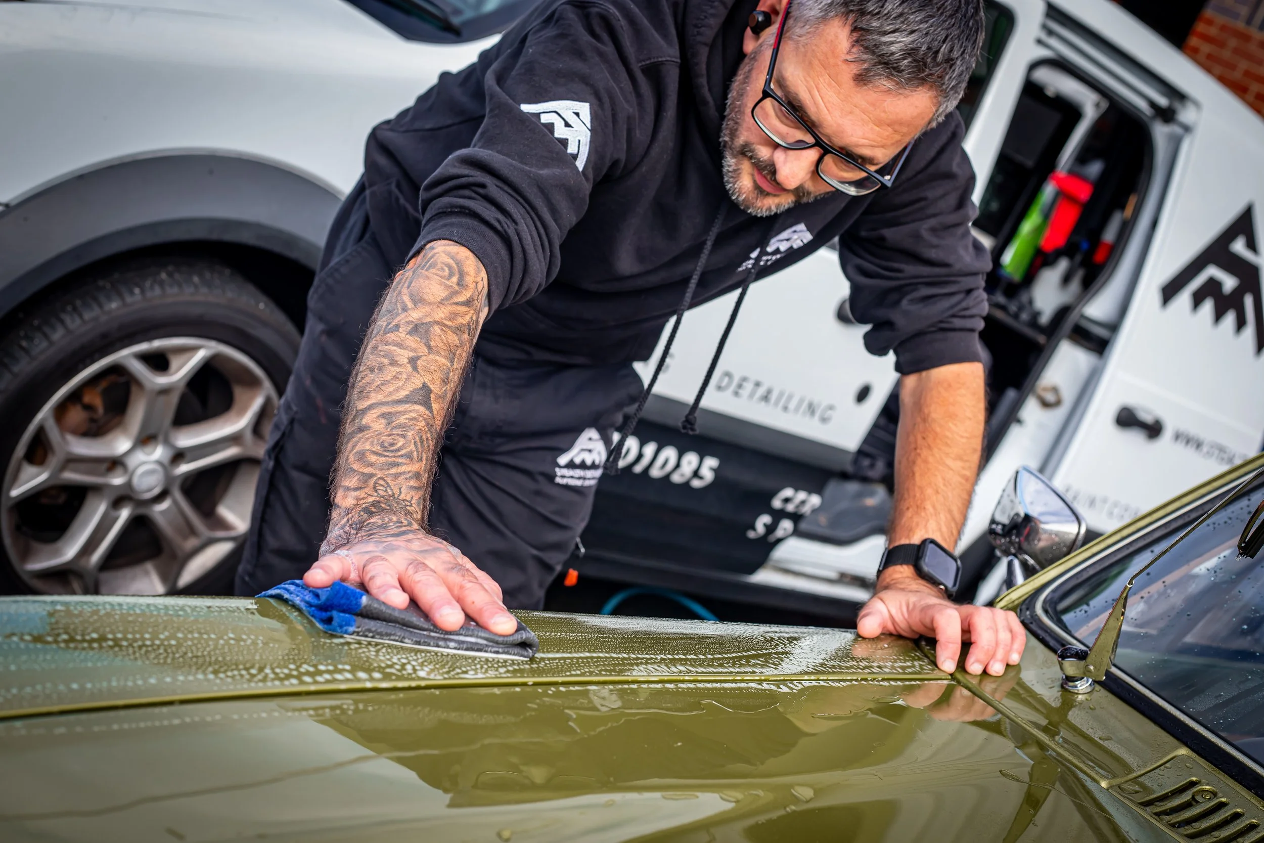 A man with tattoos and glasses cleaning a green vintage car outdoors, with a service truck in the background.